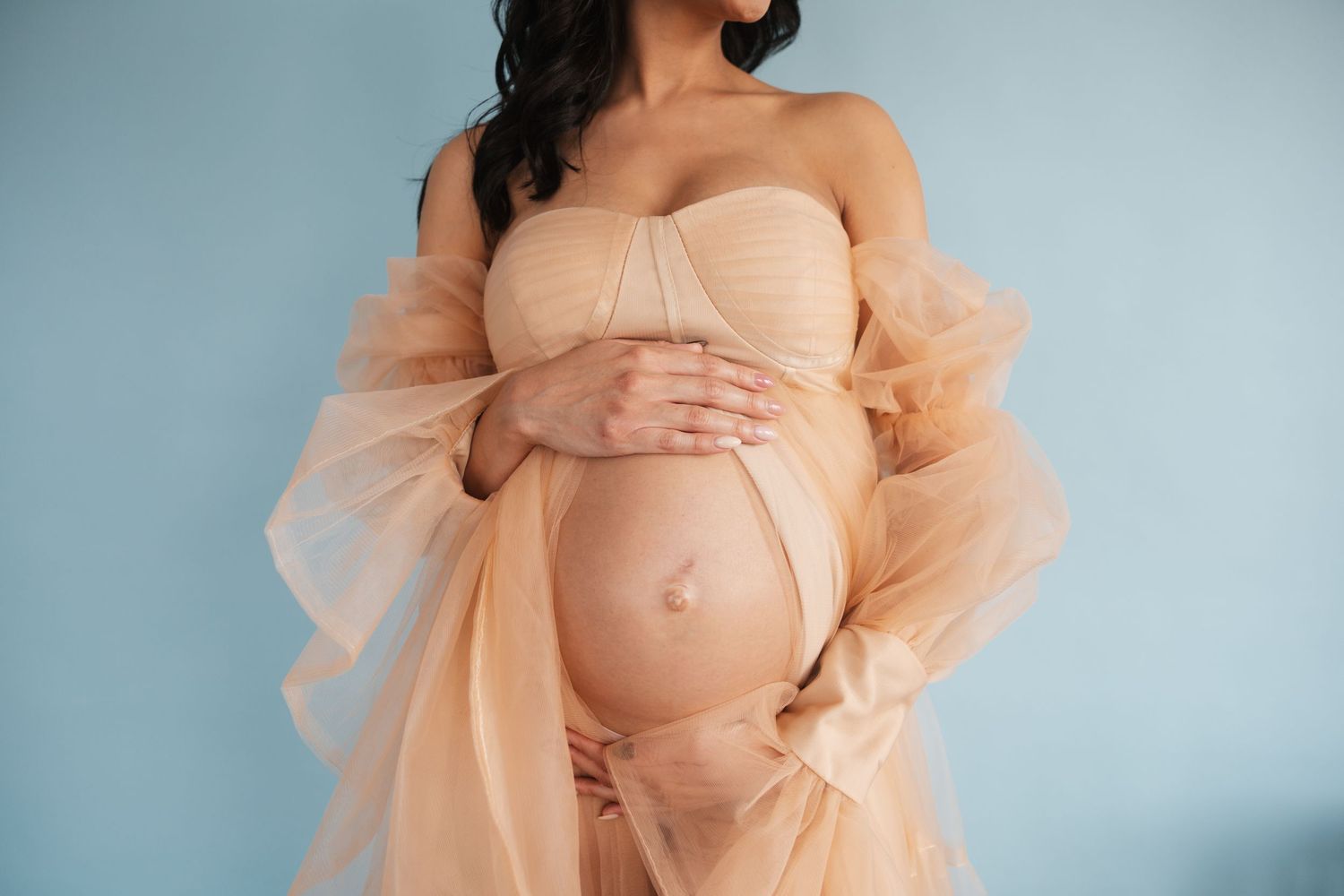 A maternity photoshoot series showing a pregnant woman in a flowing peach tulle gown against a soft blue backdrop.