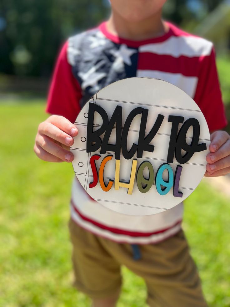 Child holding a decorative 'Back to School' sign while wearing red and white striped shirt outdoors.