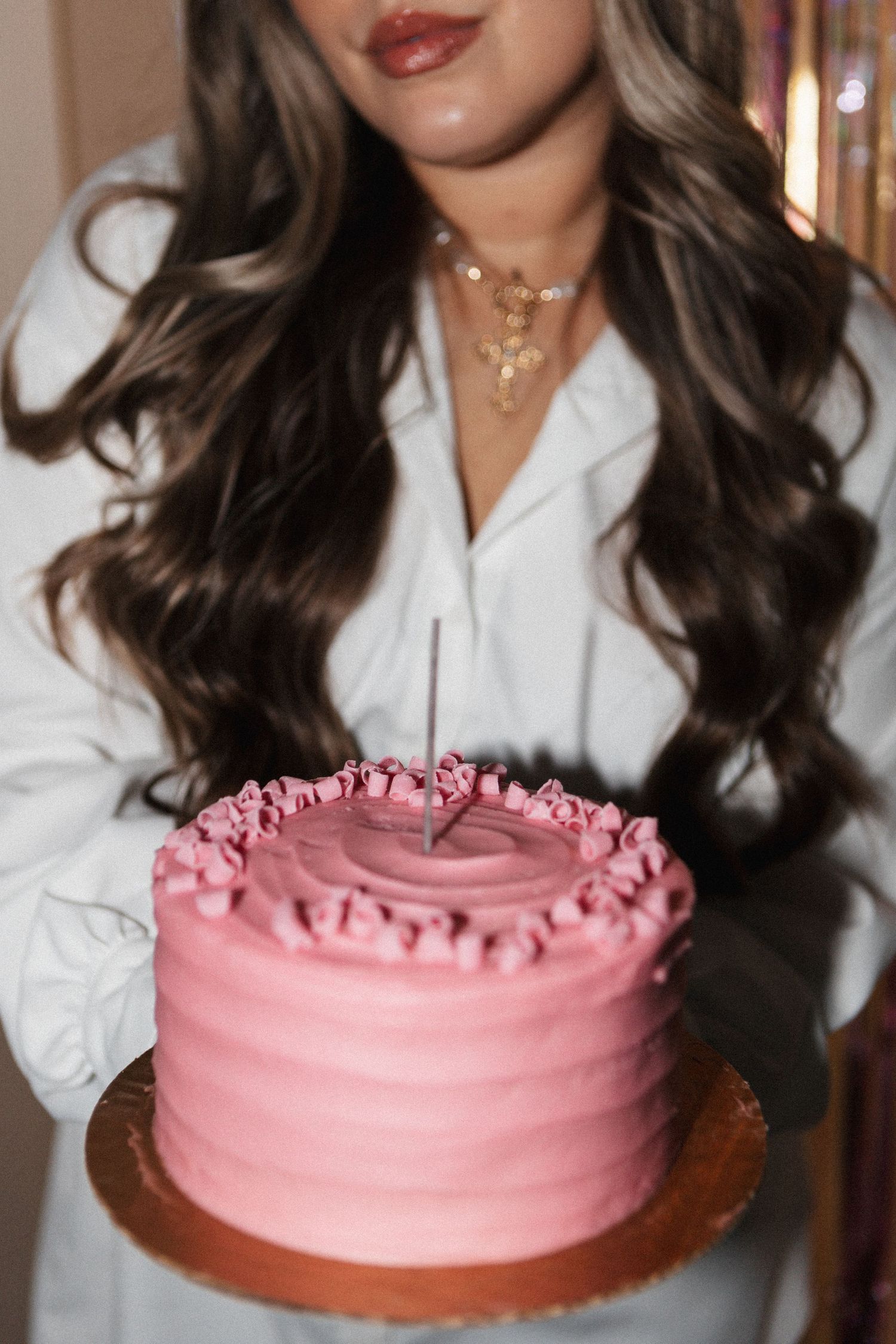 Pink frosted cake being held by someone wearing a white blouse and delicate gold necklace.