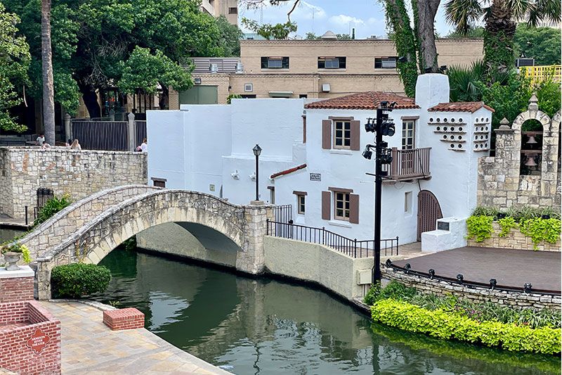 Stone bridge over calm river beside white Spanish colonial building with balcony and lush greenery in San Antonio River Walk.