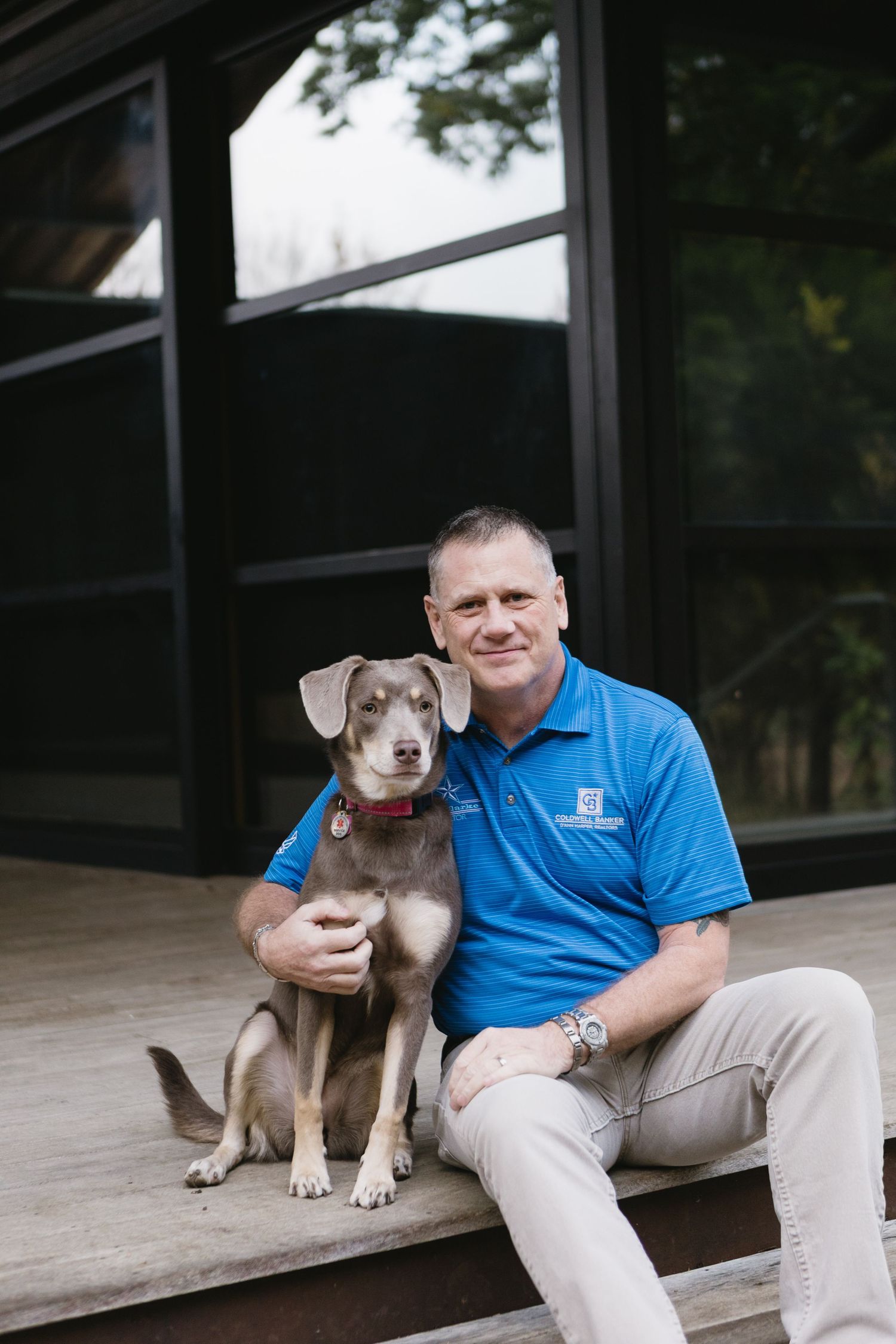 Man in blue polo shirt sitting on wooden steps with a brown dog, smiling outdoors near a modern building.