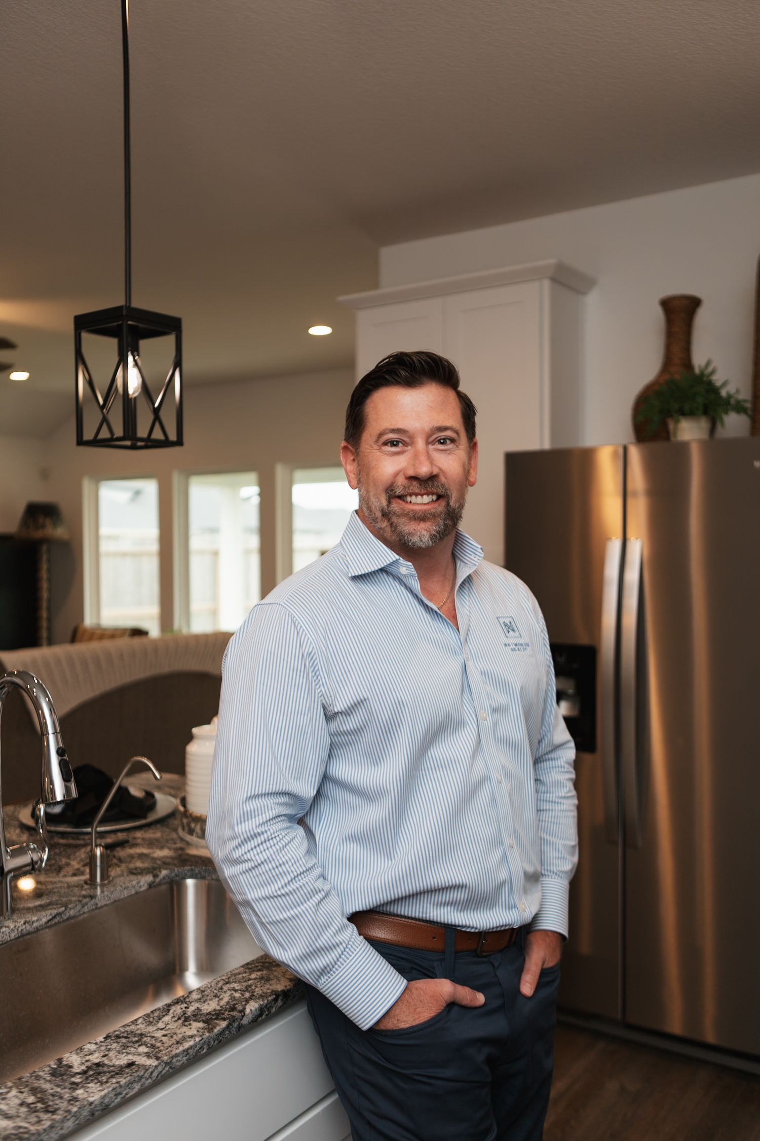 A person stands casually in a modern kitchen with granite countertops and pendant lighting.