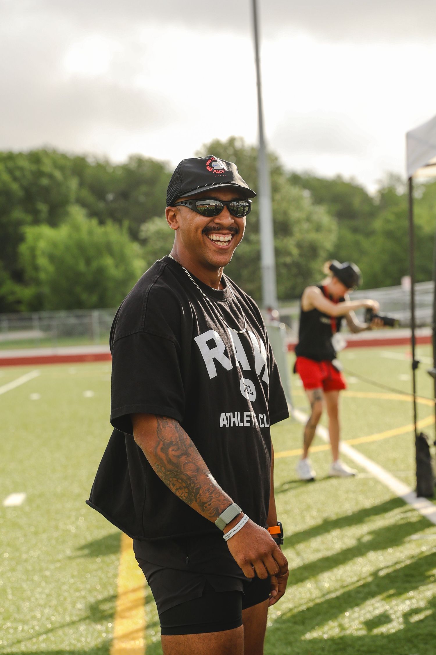 A smiling man in black RUN Athletics shirt and sunglasses stands on a sports field during an outdoor athletic event.