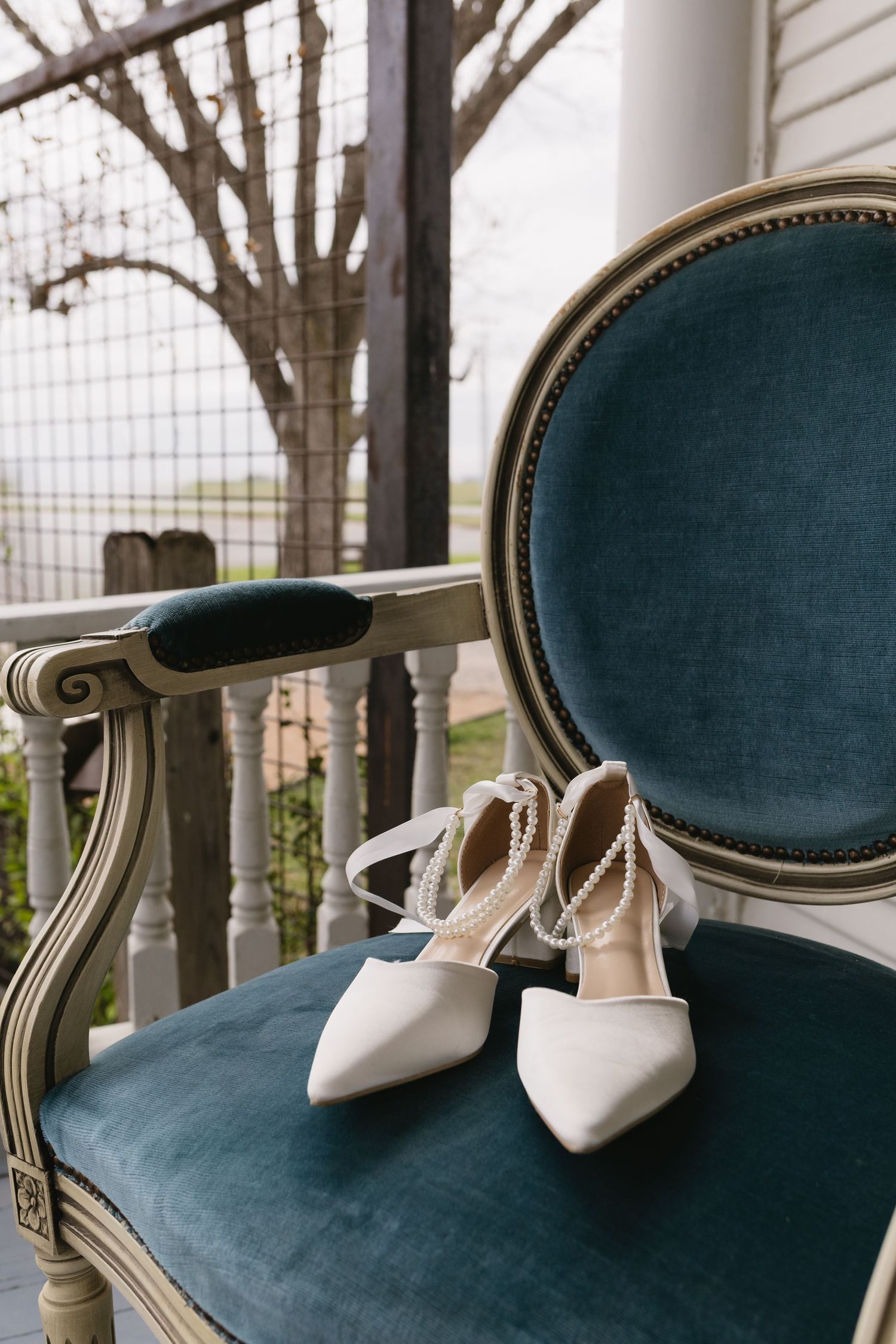 Elegant white bridal heels with pearl ankle straps resting on a teal velvet French-style chair on a porch.