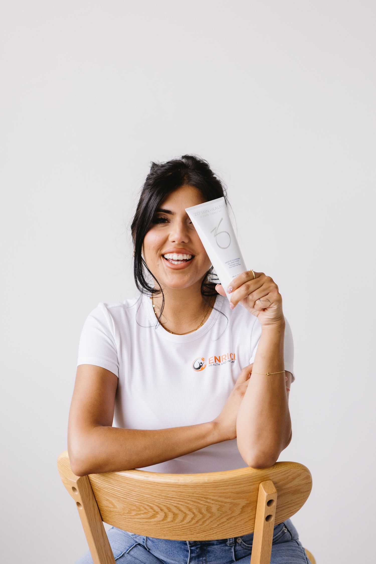 Smiling woman in white t-shirt holding skincare tube product while sitting on wooden chair against gray background.
