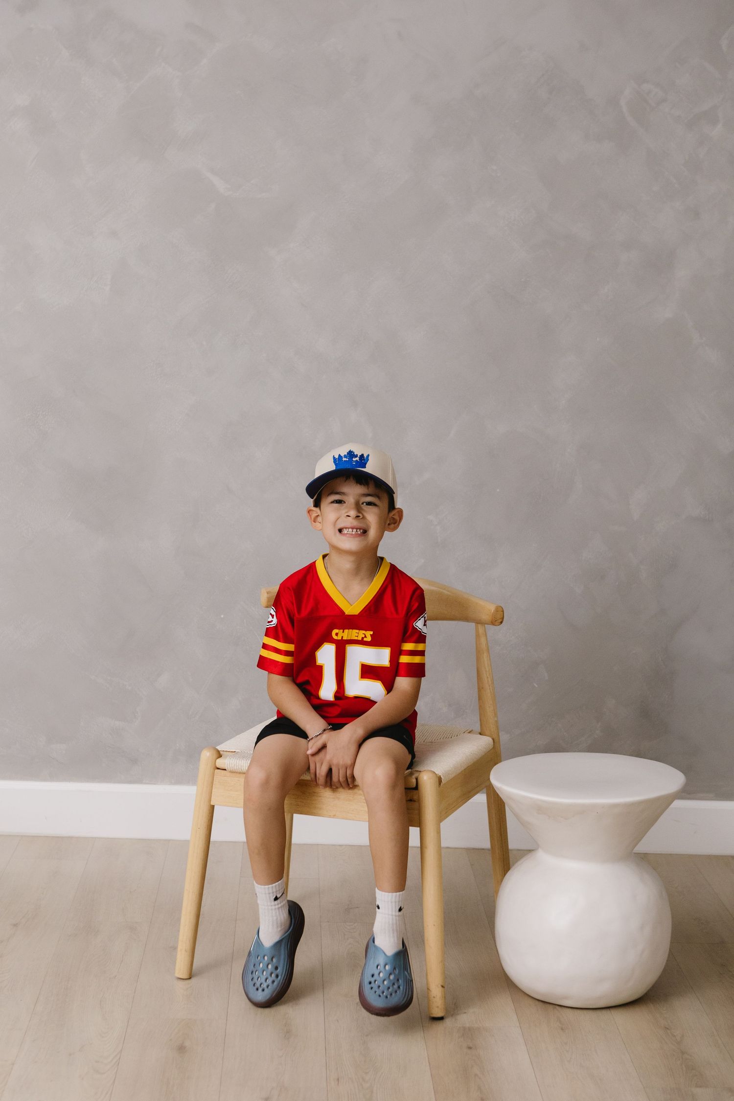 Boy in Kansas City Chiefs #15 jersey and blue cap sits smiling on wooden chair against gray wall.