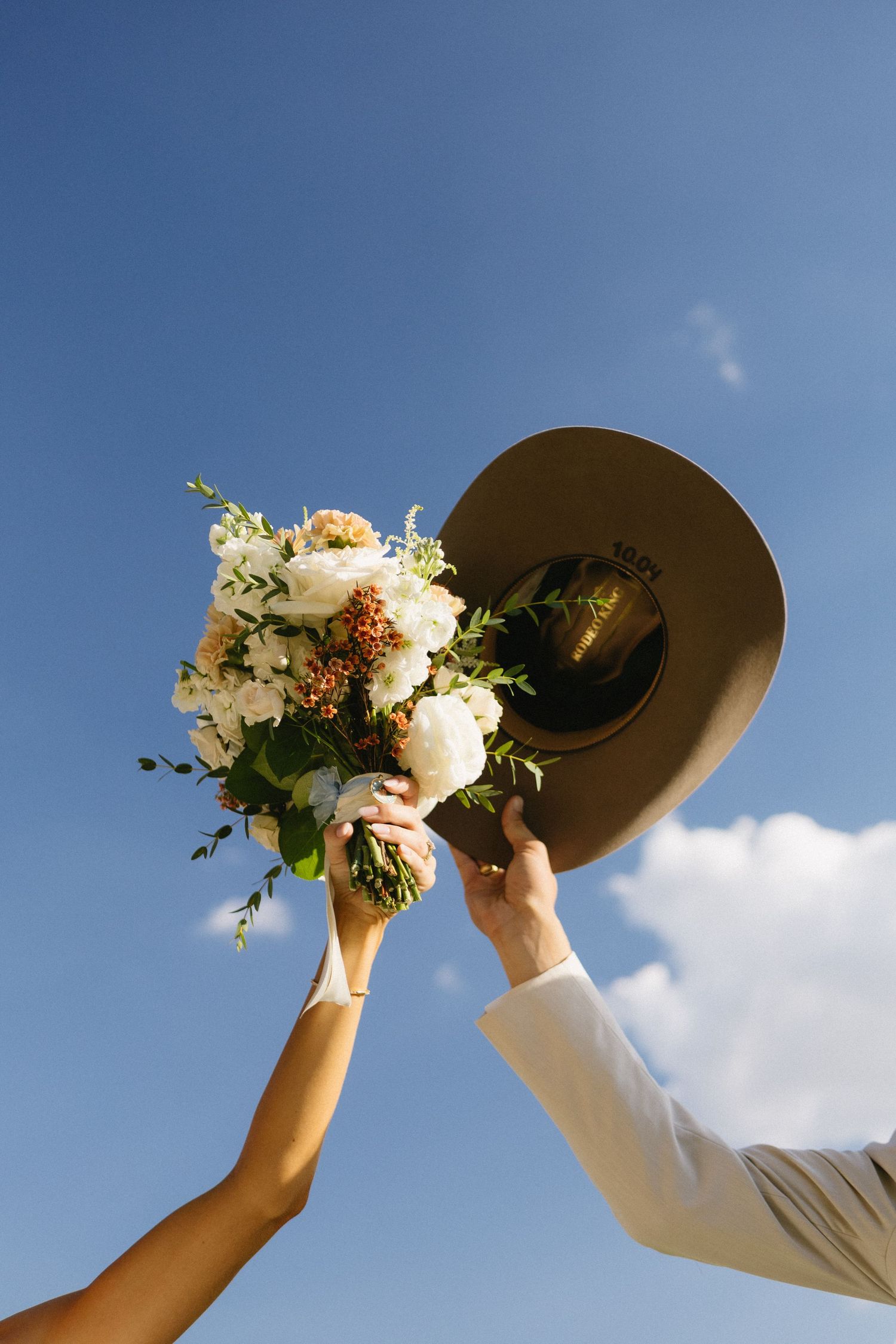 Bride and groom raising a floral bouquet and cowboy hat against a blue sky at a rustic outdoor wedding.