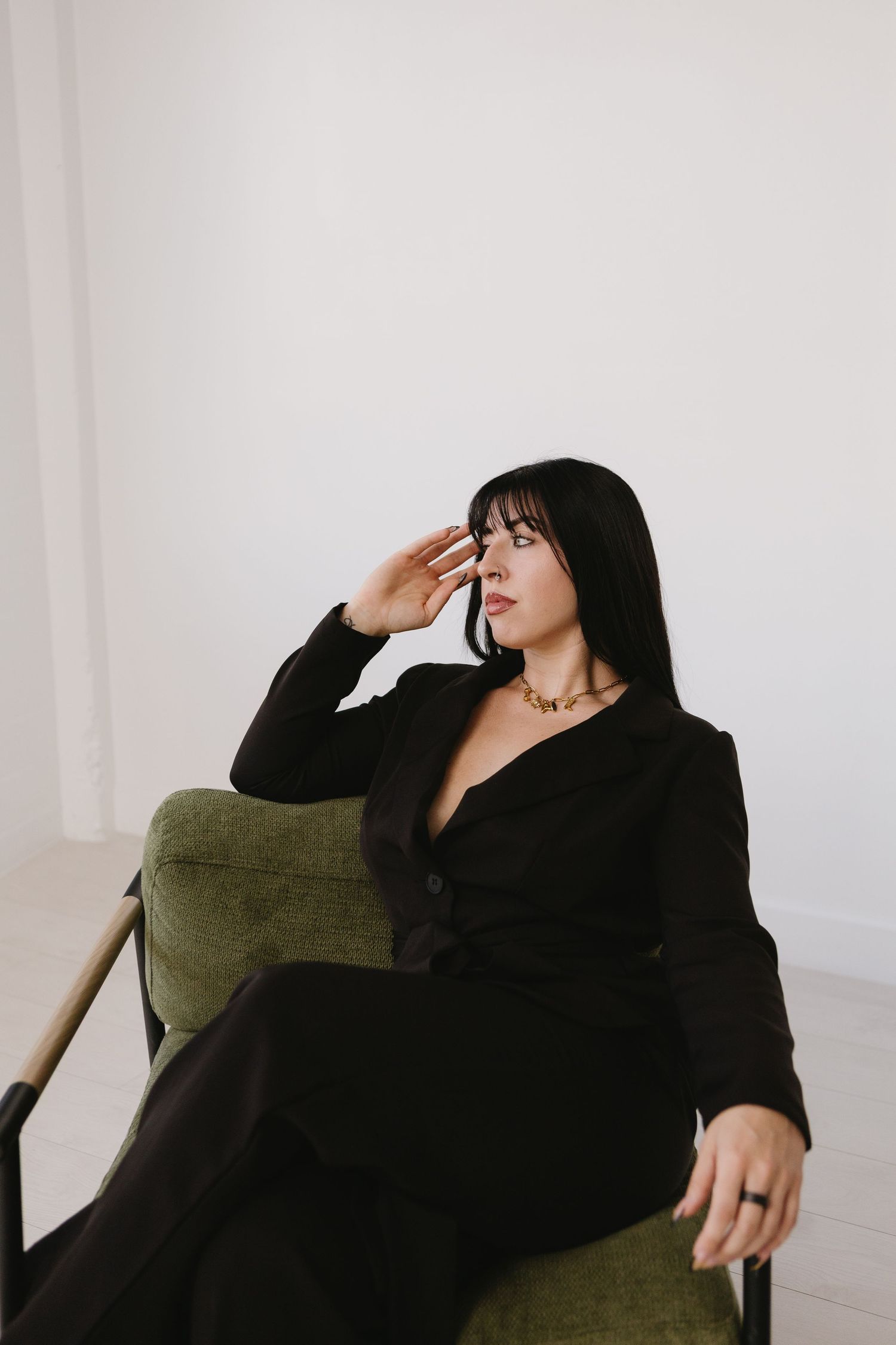 Woman in black blazer suit seated in green chair shielding eyes with hand against white background.