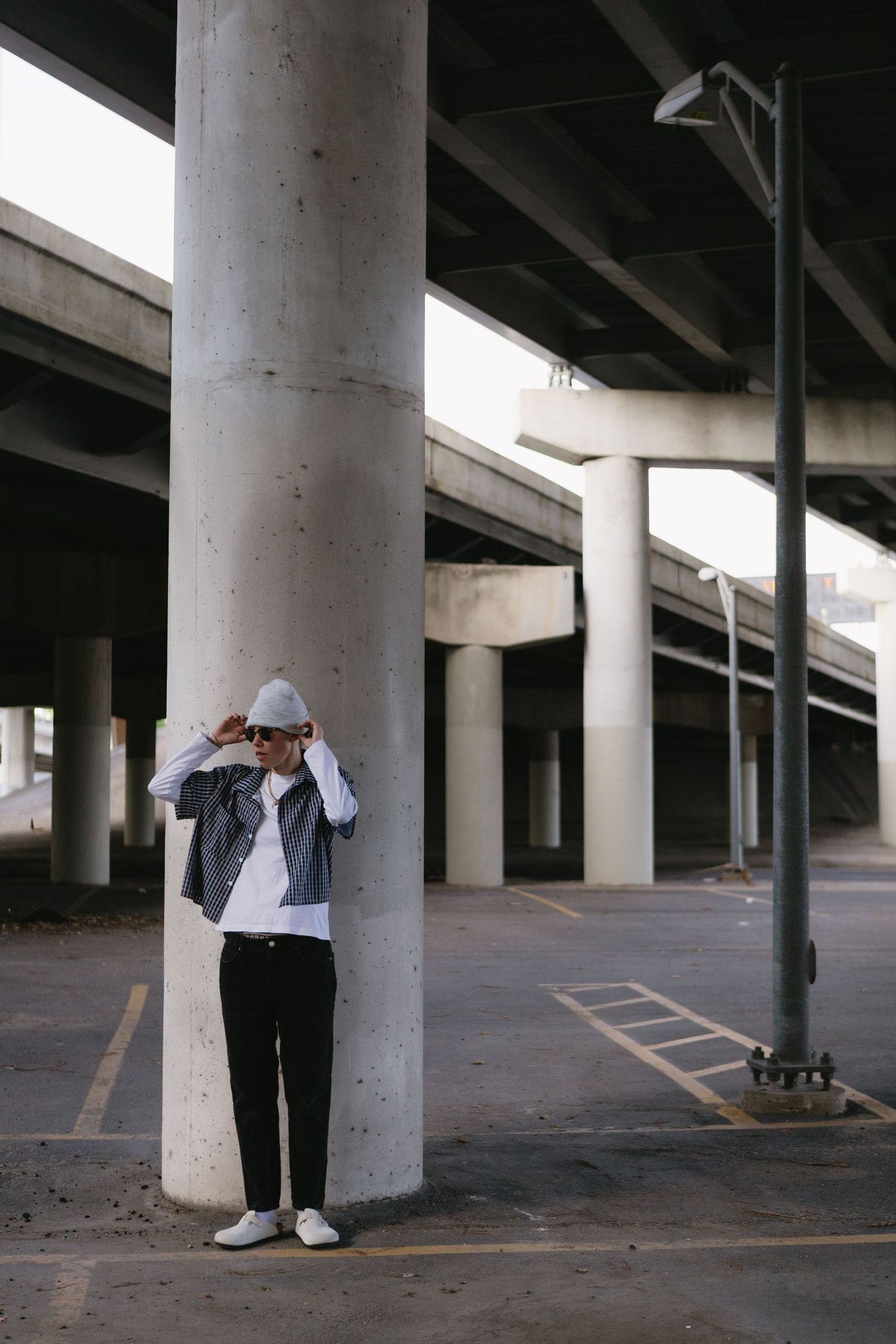 Young man in gray beanie, denim jacket, and black pants leaning against concrete pillar under urban highway overpass.