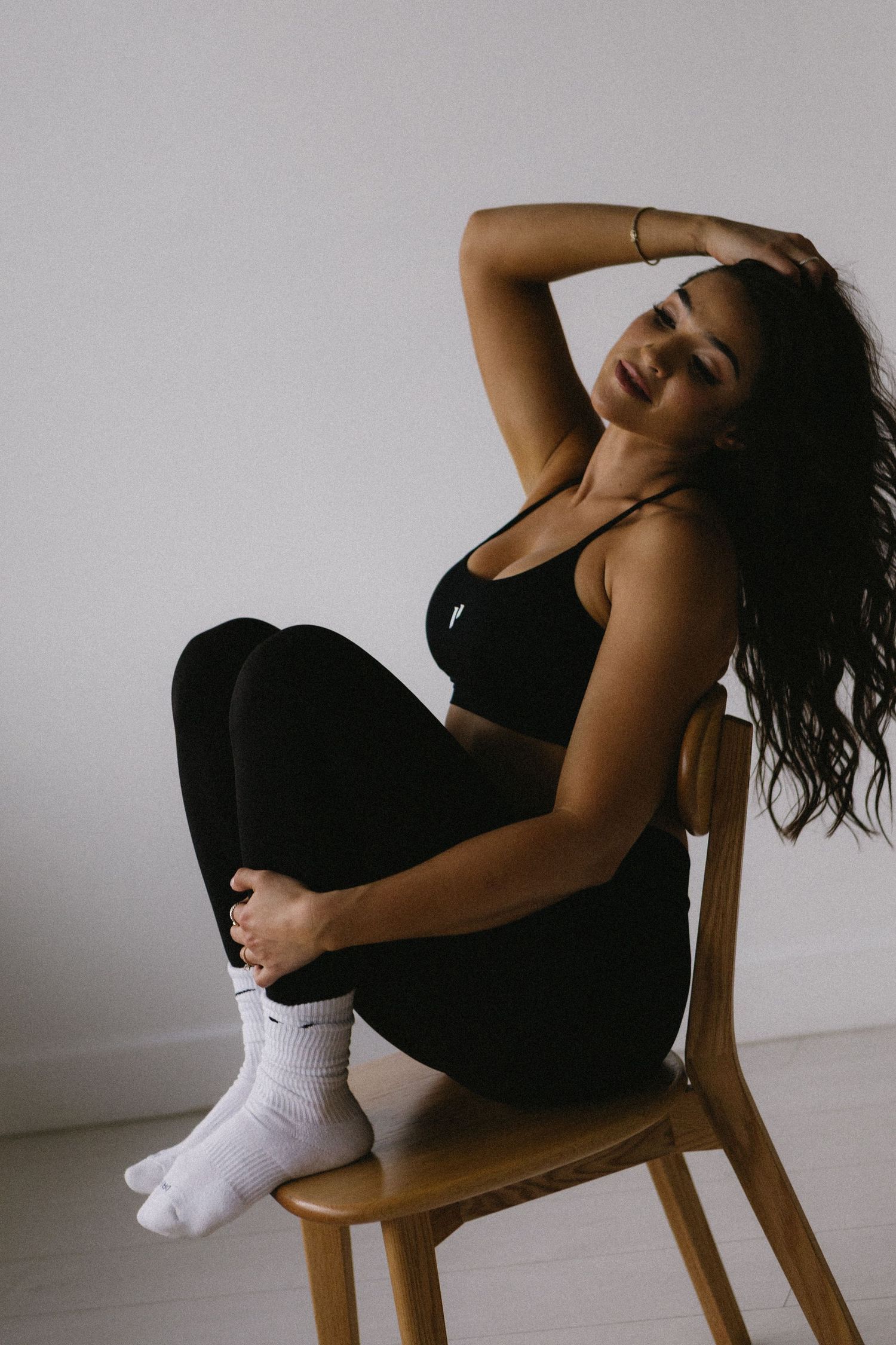 Woman in black sports bra and leggings with white Nike socks poses on wooden chair against white wall.