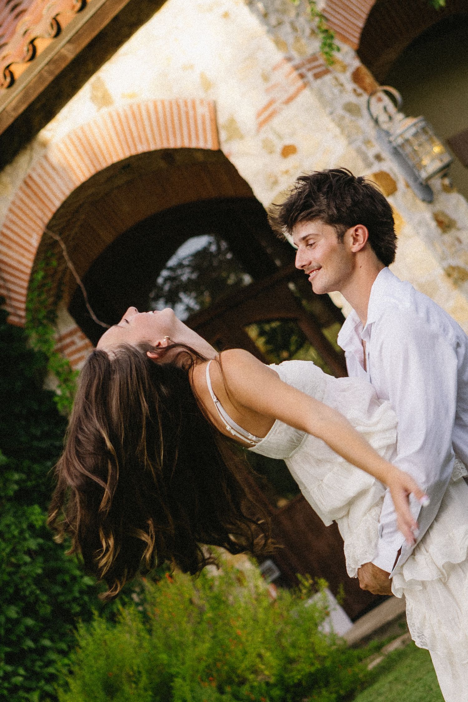 A joyful couple in white attire dancing and spinning outdoors near rustic stone arches surrounded by lush greenery.