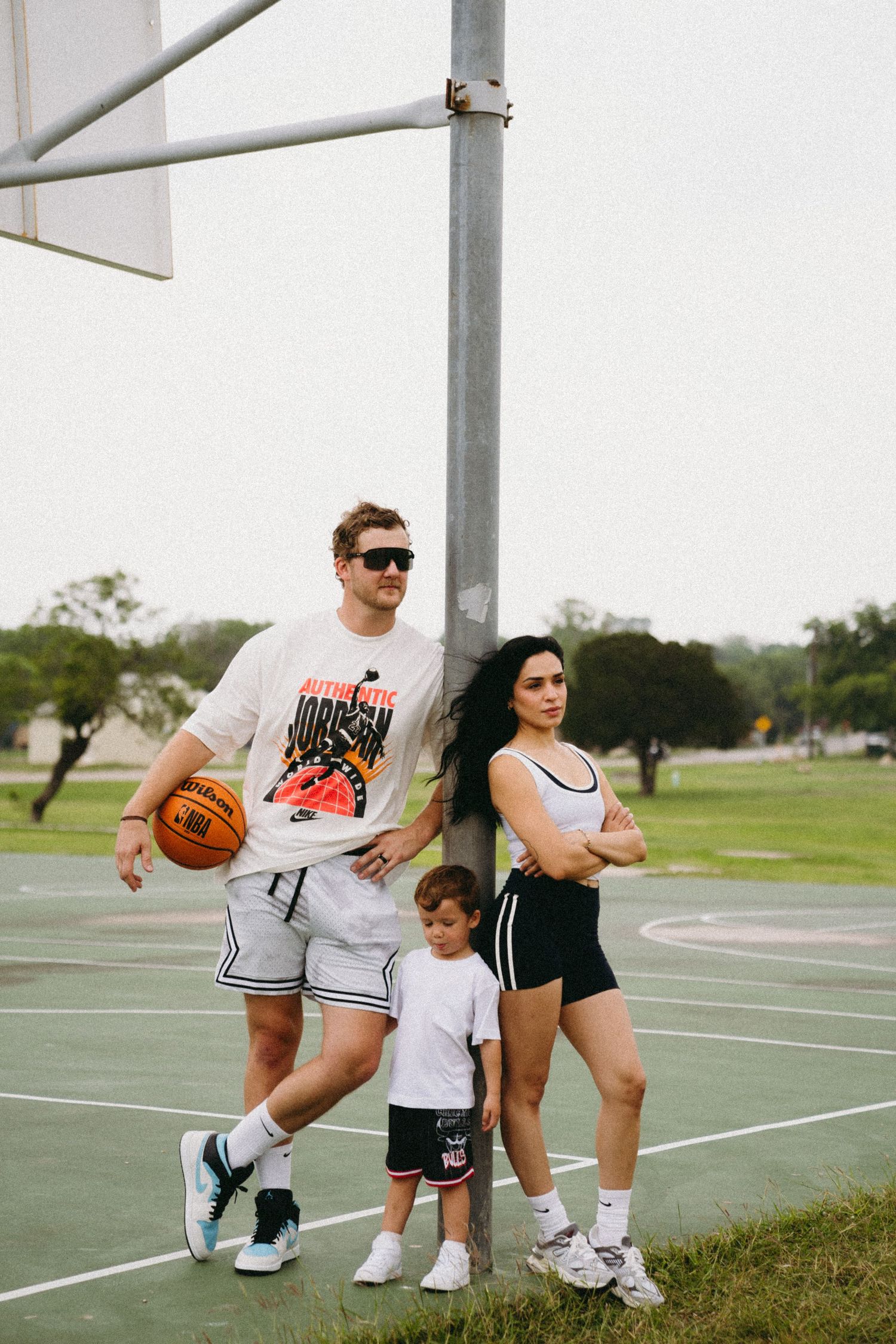 Family of three posing on outdoor basketball court, man holding ball, woman with crossed arms, child standing between them.