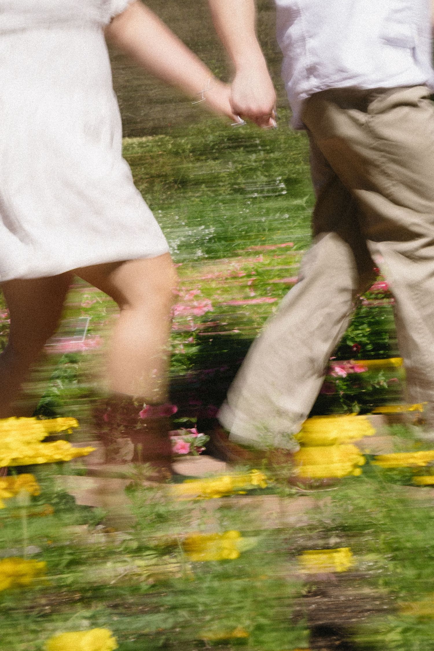 Blurred motion shot of a couple holding hands walking through a colorful garden with yellow and pink flowers.
