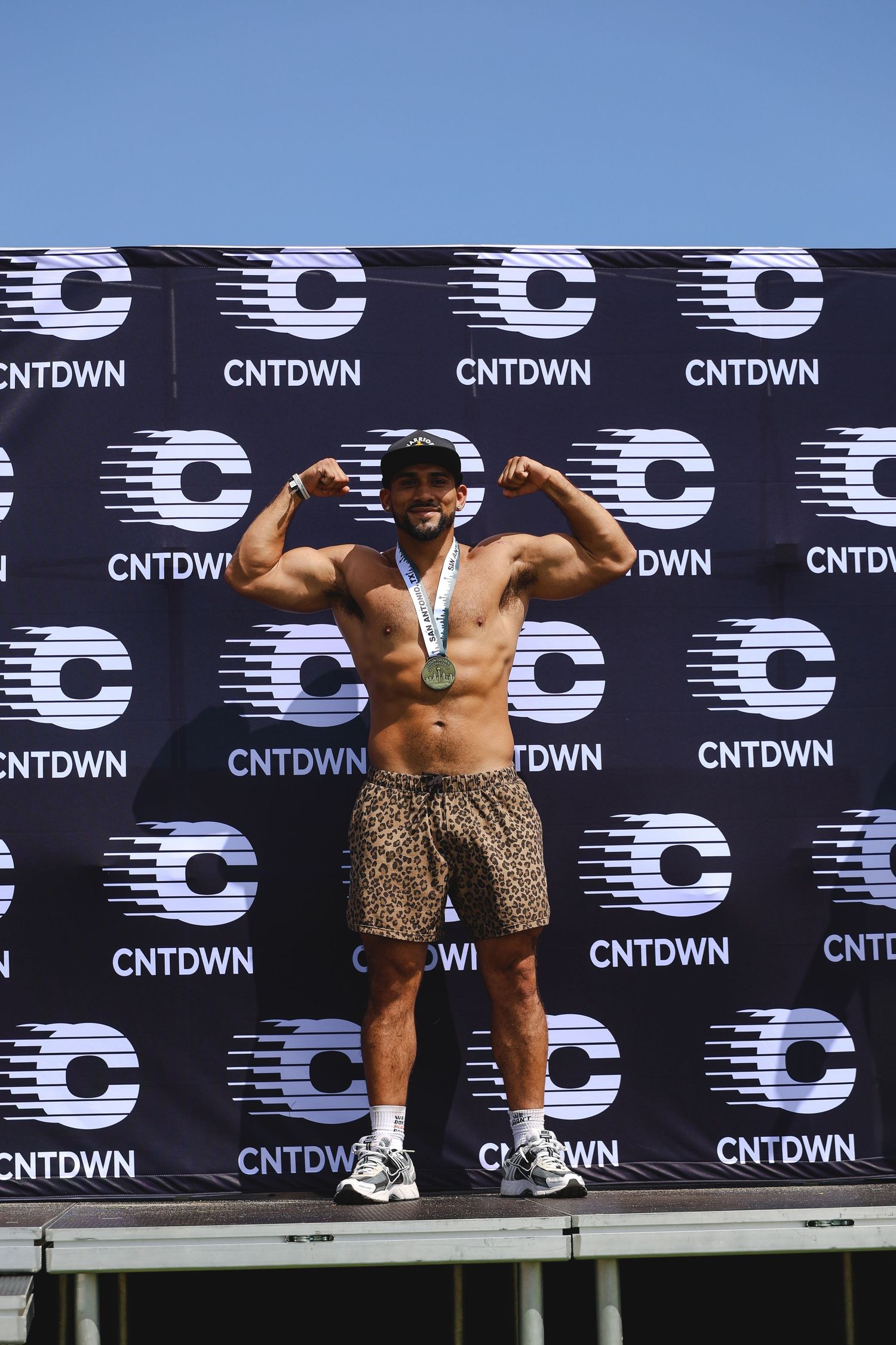 Muscular man flexing biceps on podium wearing medal and leopard print shorts at CNTDWN event.
