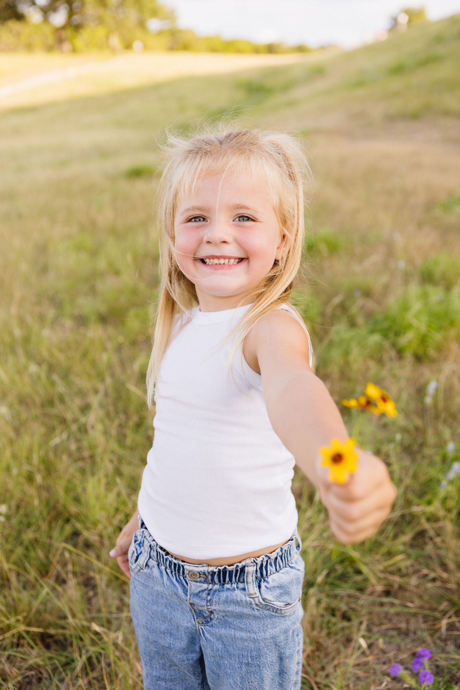 Smiling blonde girl in white tank top and jeans holding a yellow wildflower in a sunny green meadow.