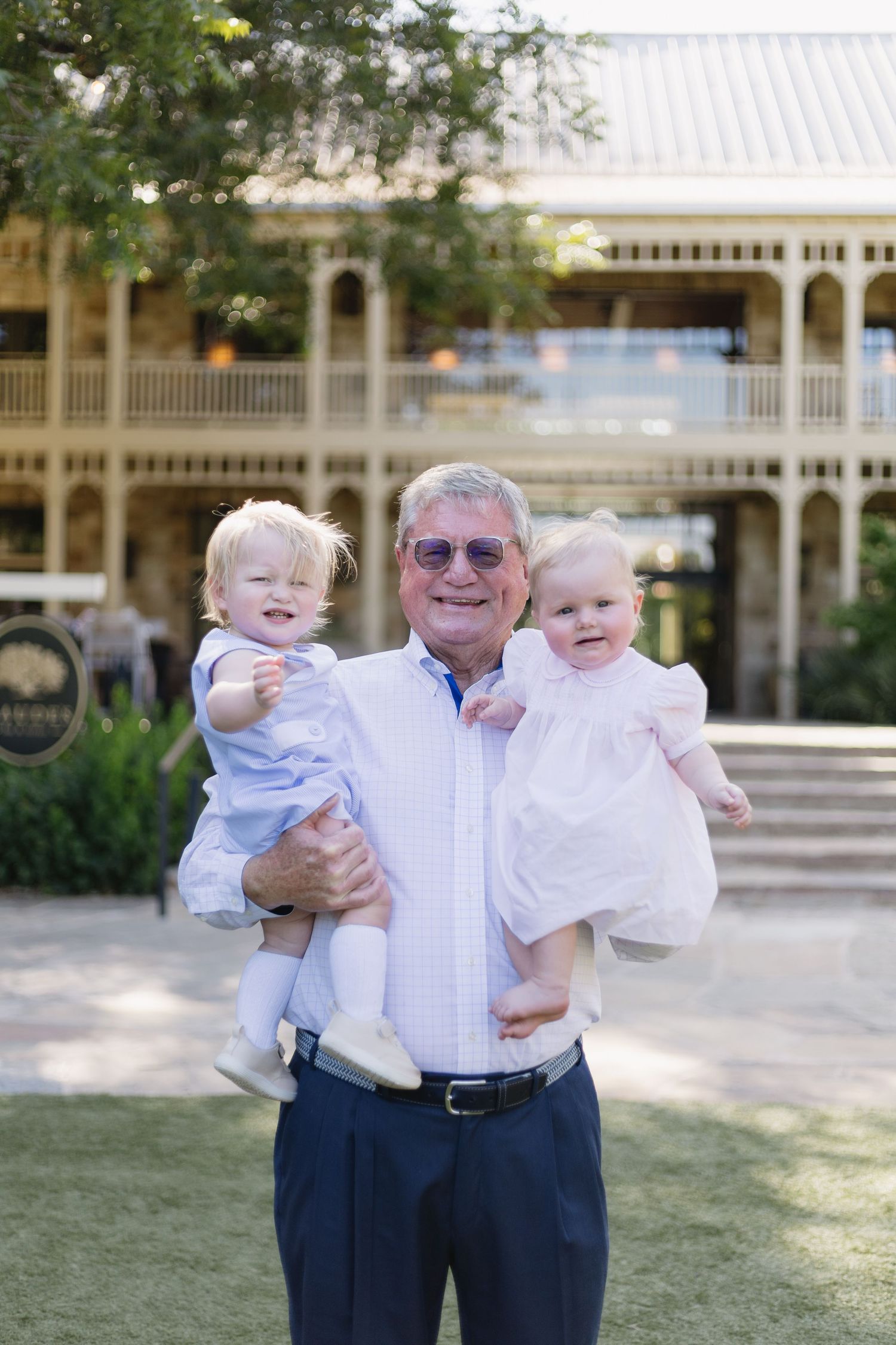 Elderly man in sunglasses holding two toddlers outdoors near a colonial-style building on a sunny day.