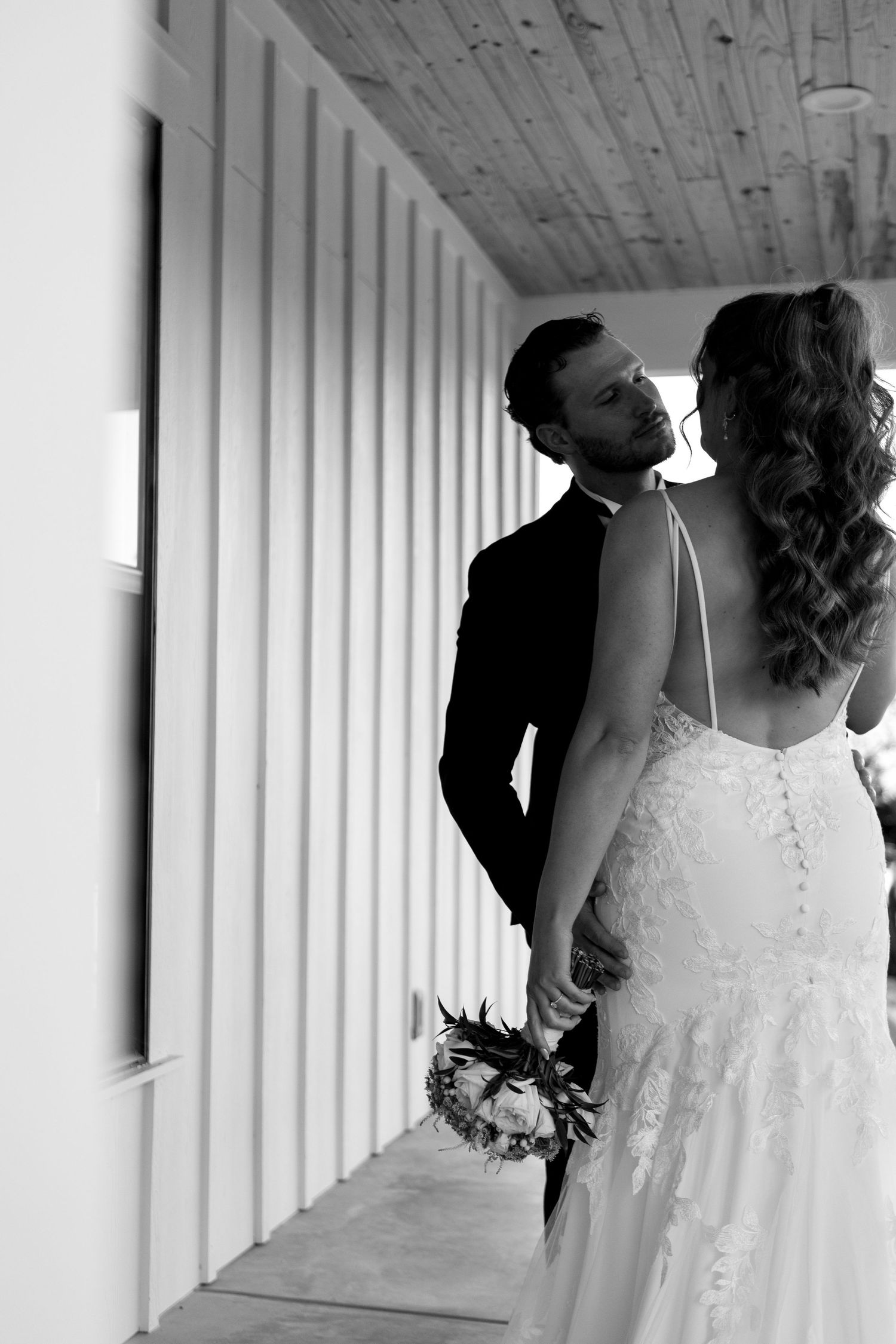 Black and white photo of bride and groom holding hands, sharing a romantic glance on a white wooden porch.