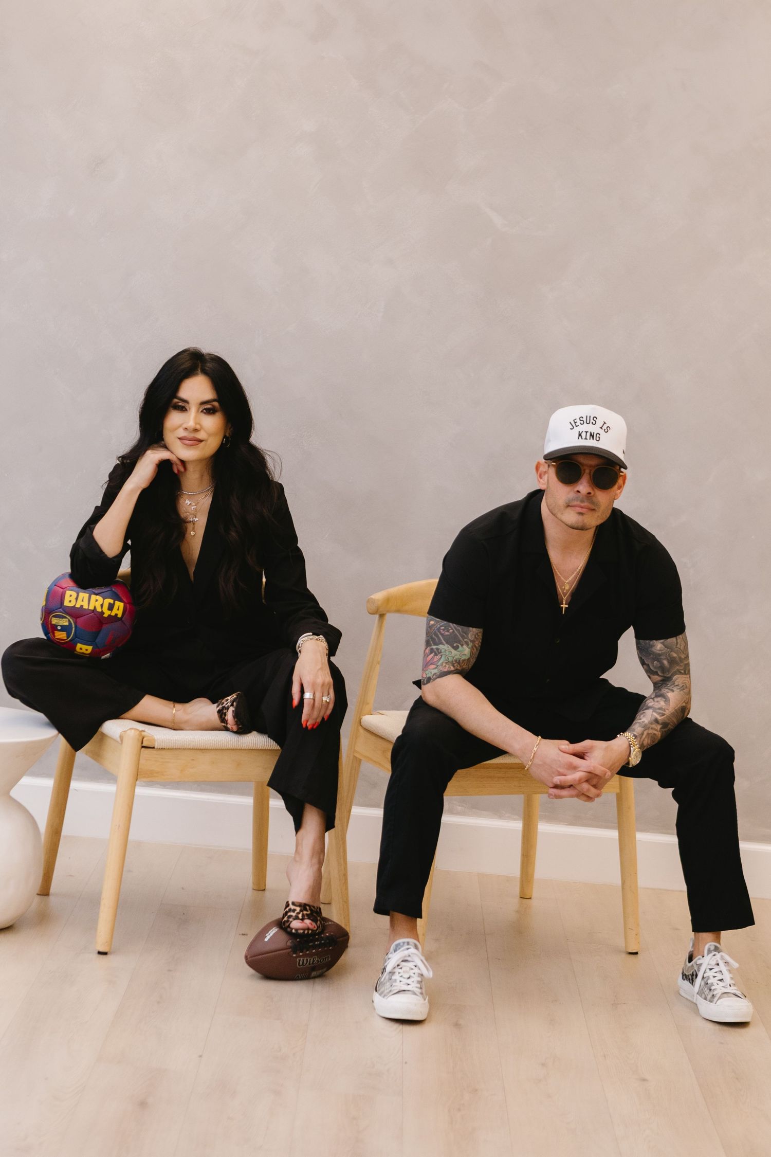 Two stylish people in black outfits sit on wooden chairs against a gray wall, one holding a FC Barcelona bag.