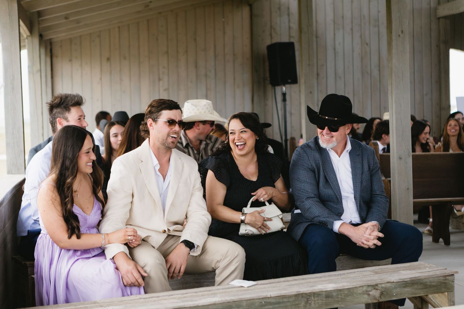 Guests smile and laugh while seated at an outdoor rustic wooden venue during a country-themed wedding ceremony.