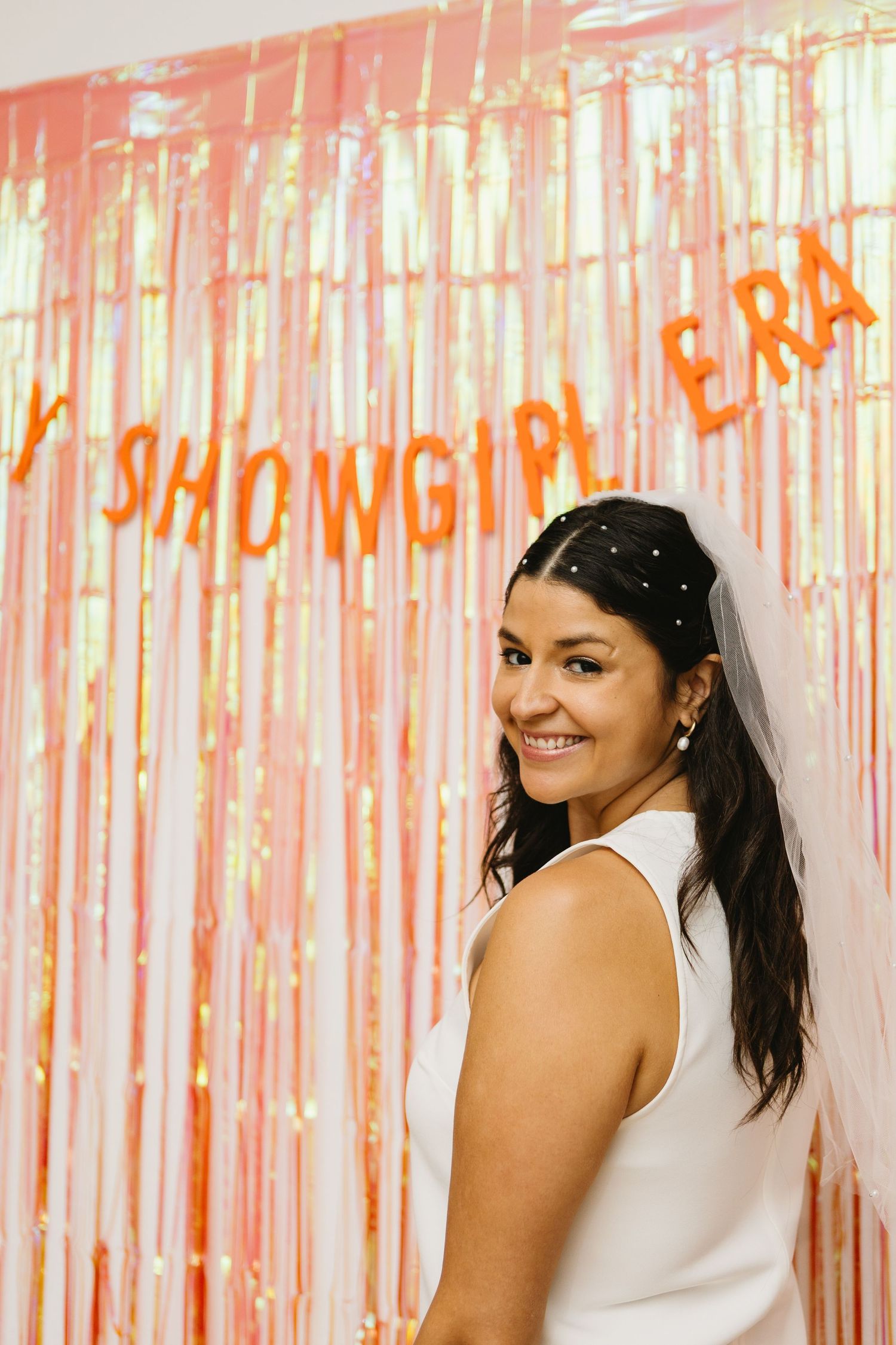 Smiling bride in white dress poses before a rose gold tinsel backdrop with orange balloon letters reading MY SHOWGIRL ERA.