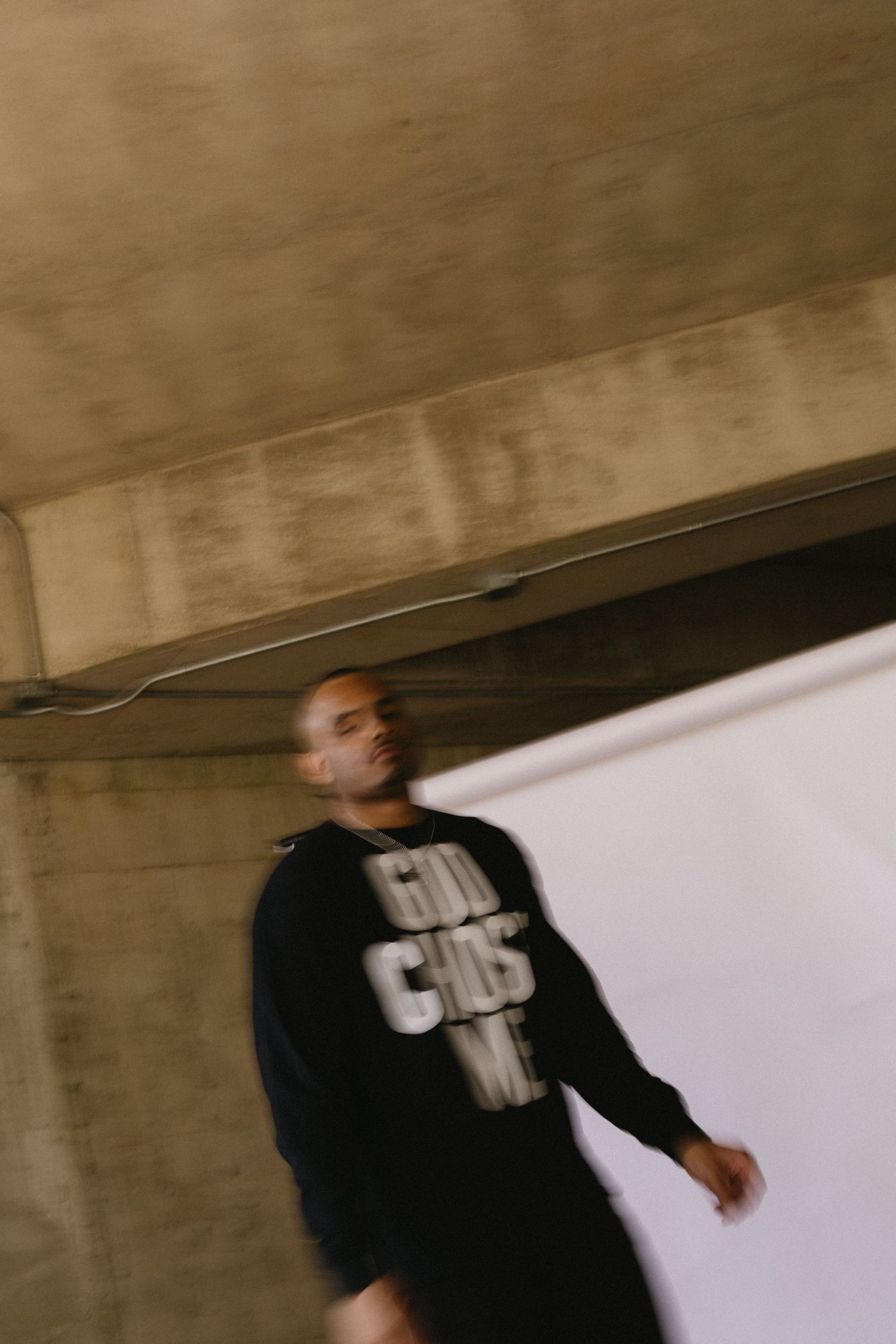 Man in black hoodie with graphic text stands in blurred motion under concrete structure in moody urban setting.