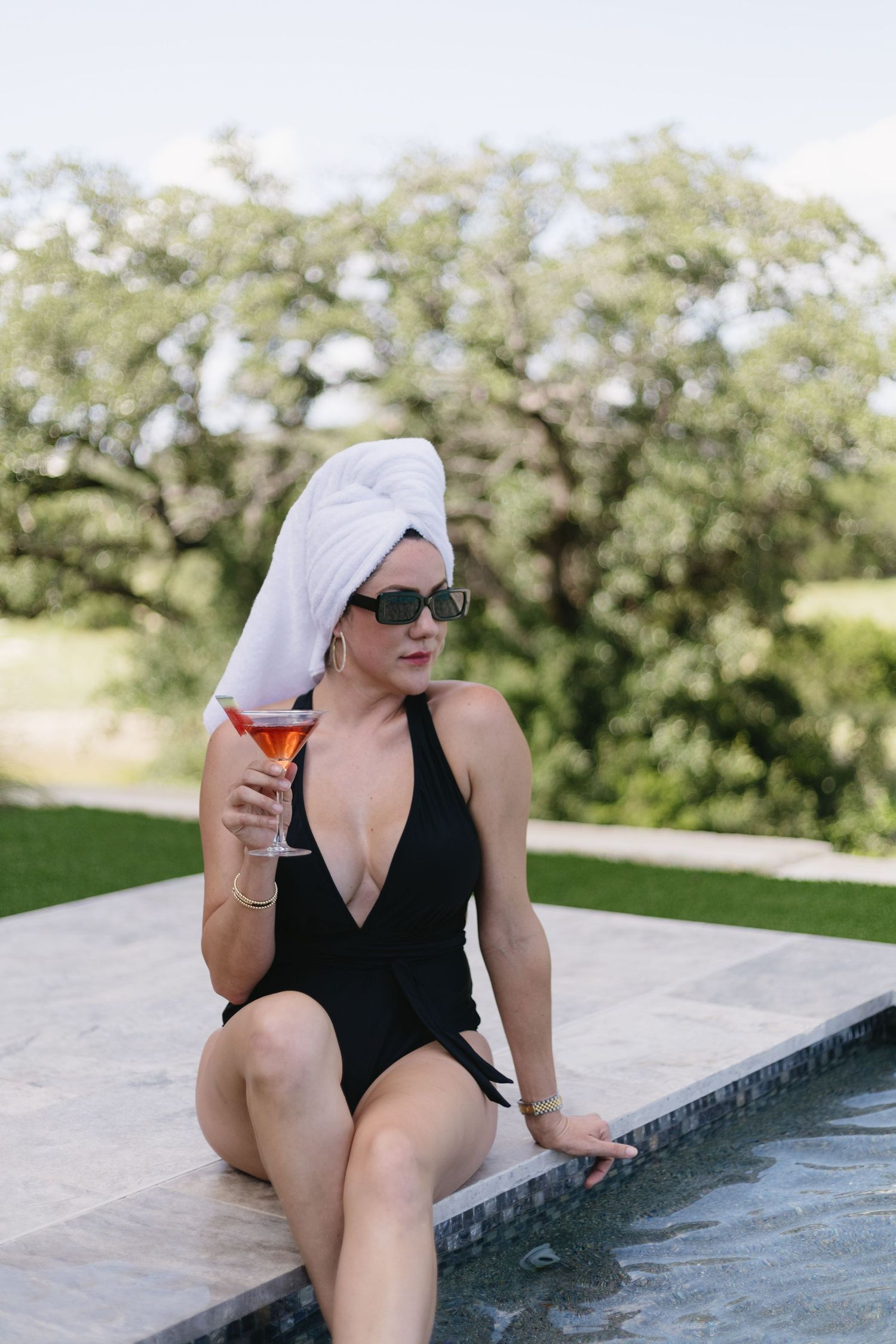 Elegant woman in black swimsuit and hair towel sips cocktail by infinity pool with scenic view.
