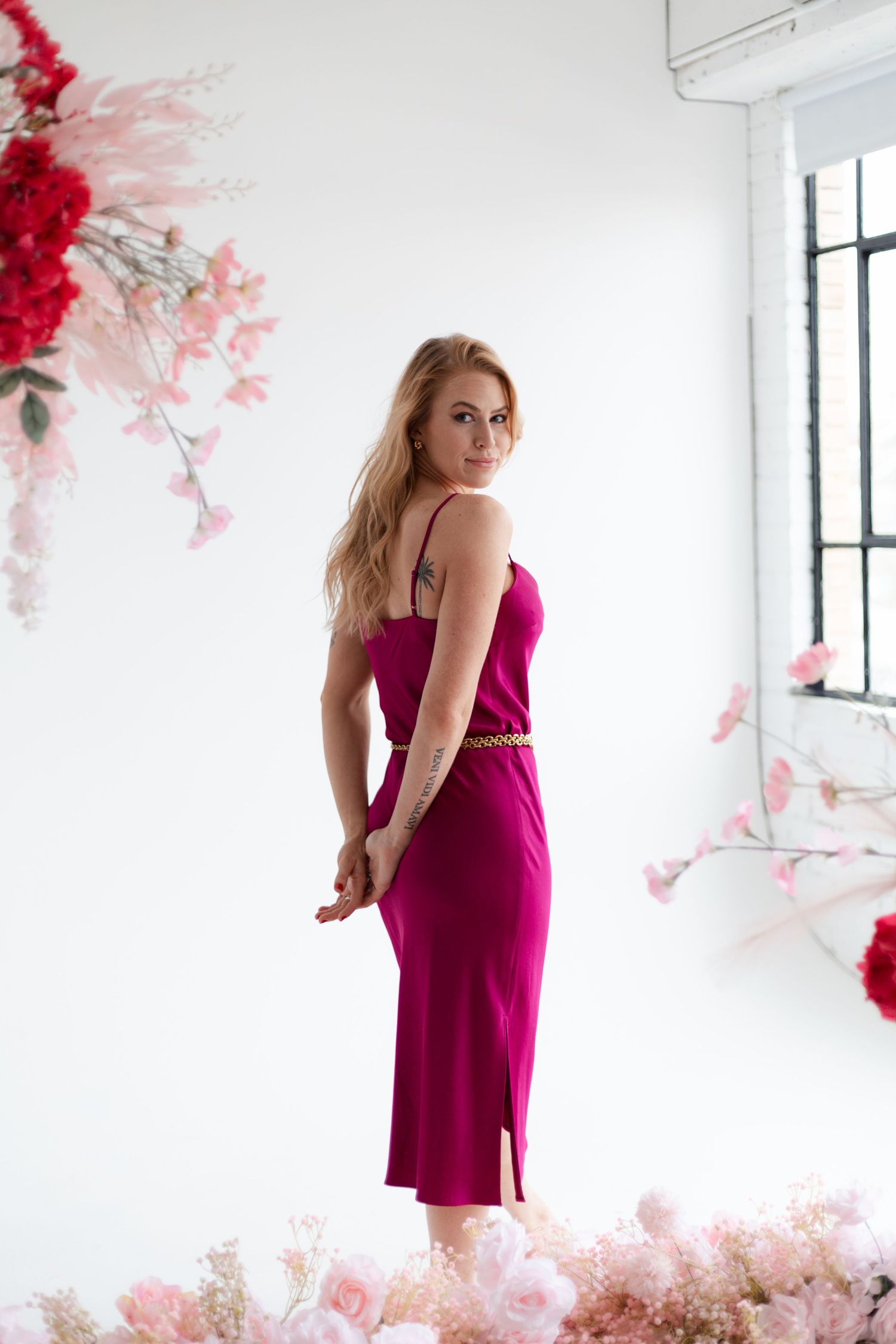 Person in magenta dress poses elegantly in white room with pink floral accents.