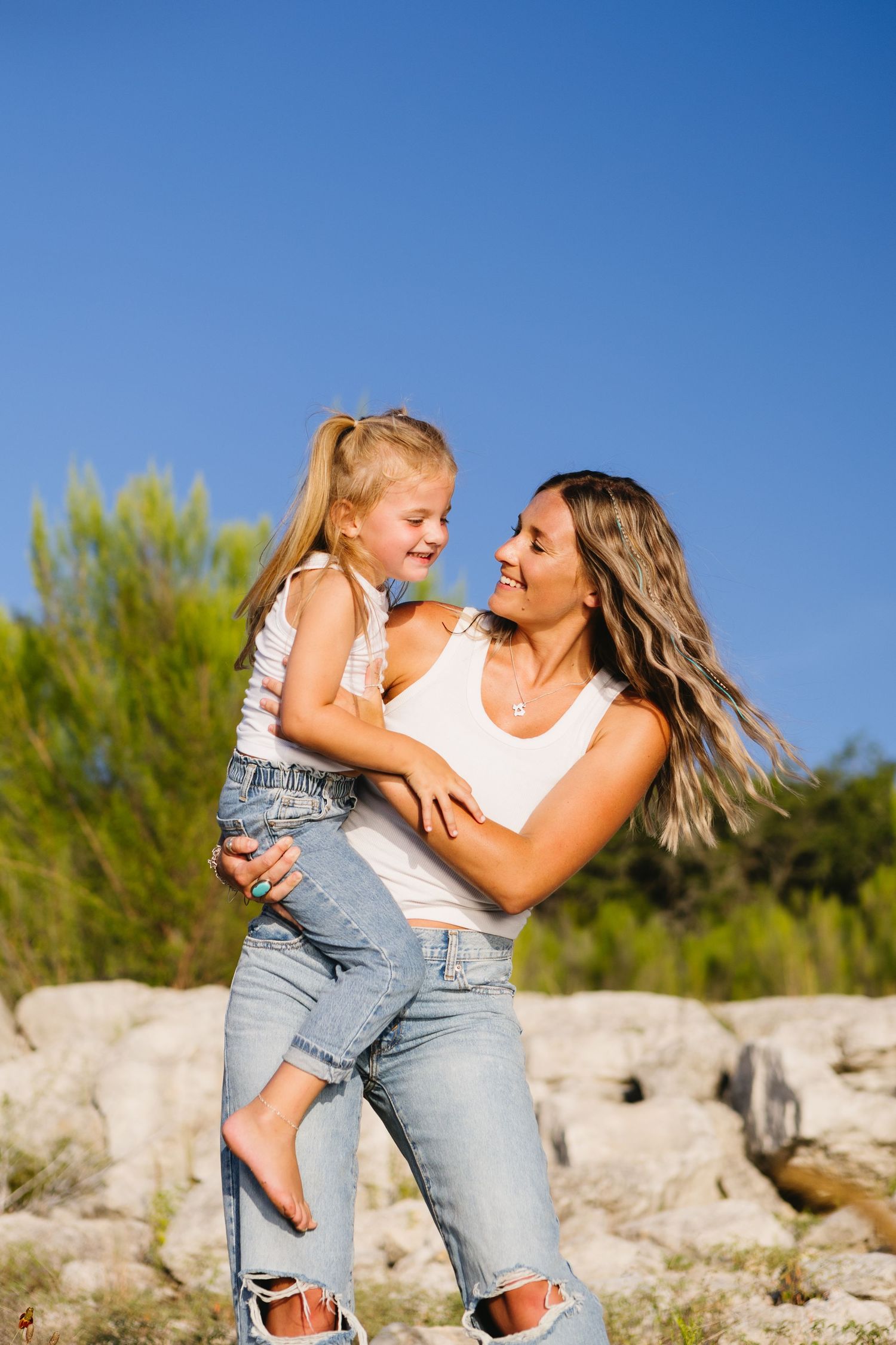 A smiling mother holds her laughing young daughter outdoors, both wearing white tops and jeans against a blue sky.