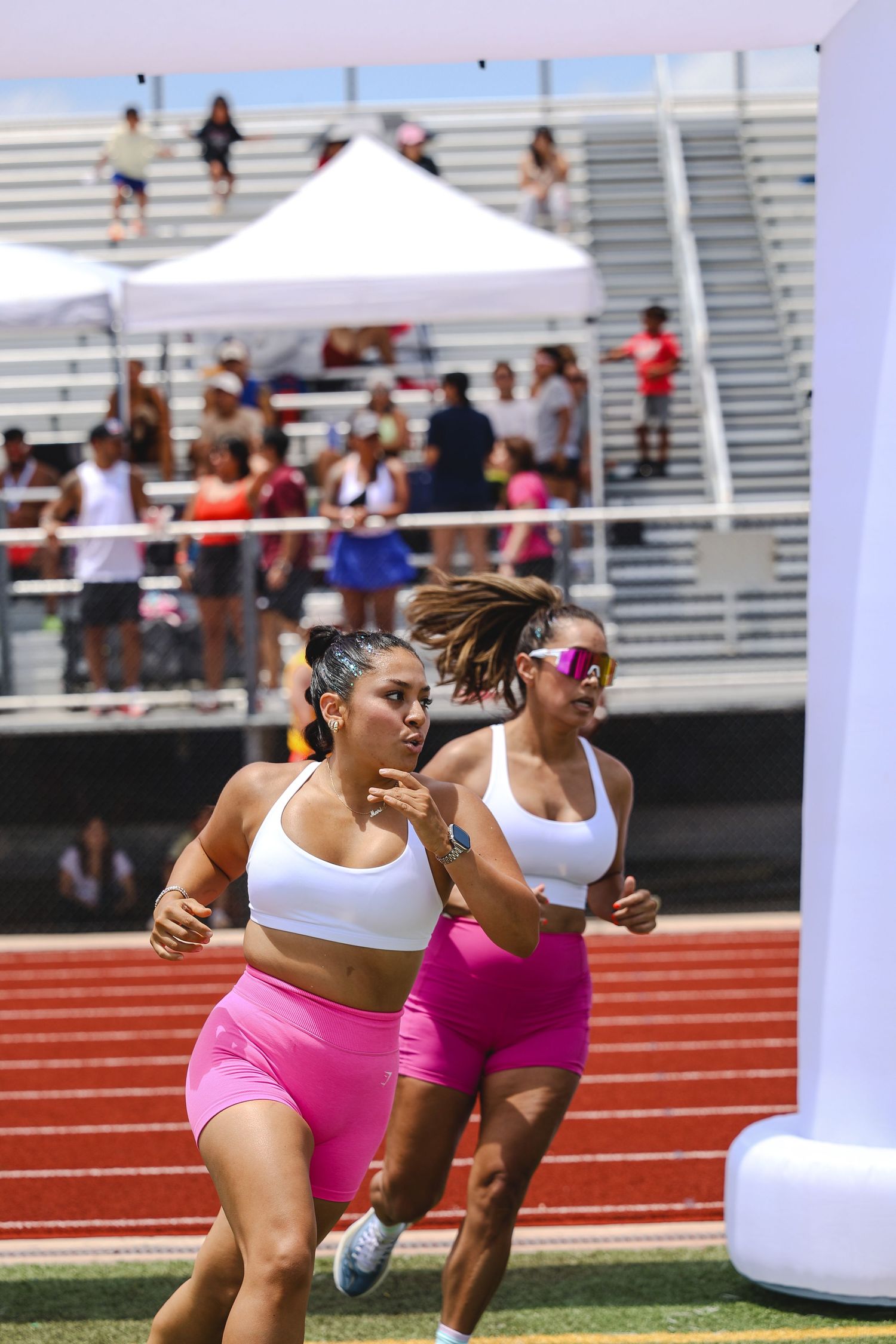 Two women in matching pink shorts and white sports bras sprint on a red track at an outdoor athletic competition.