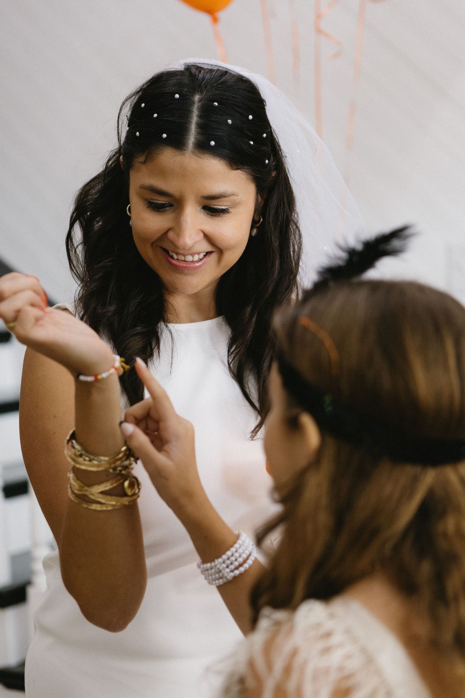 Smiling bride in white dress and veil admires gold bracelets with a guest at a wedding celebration.