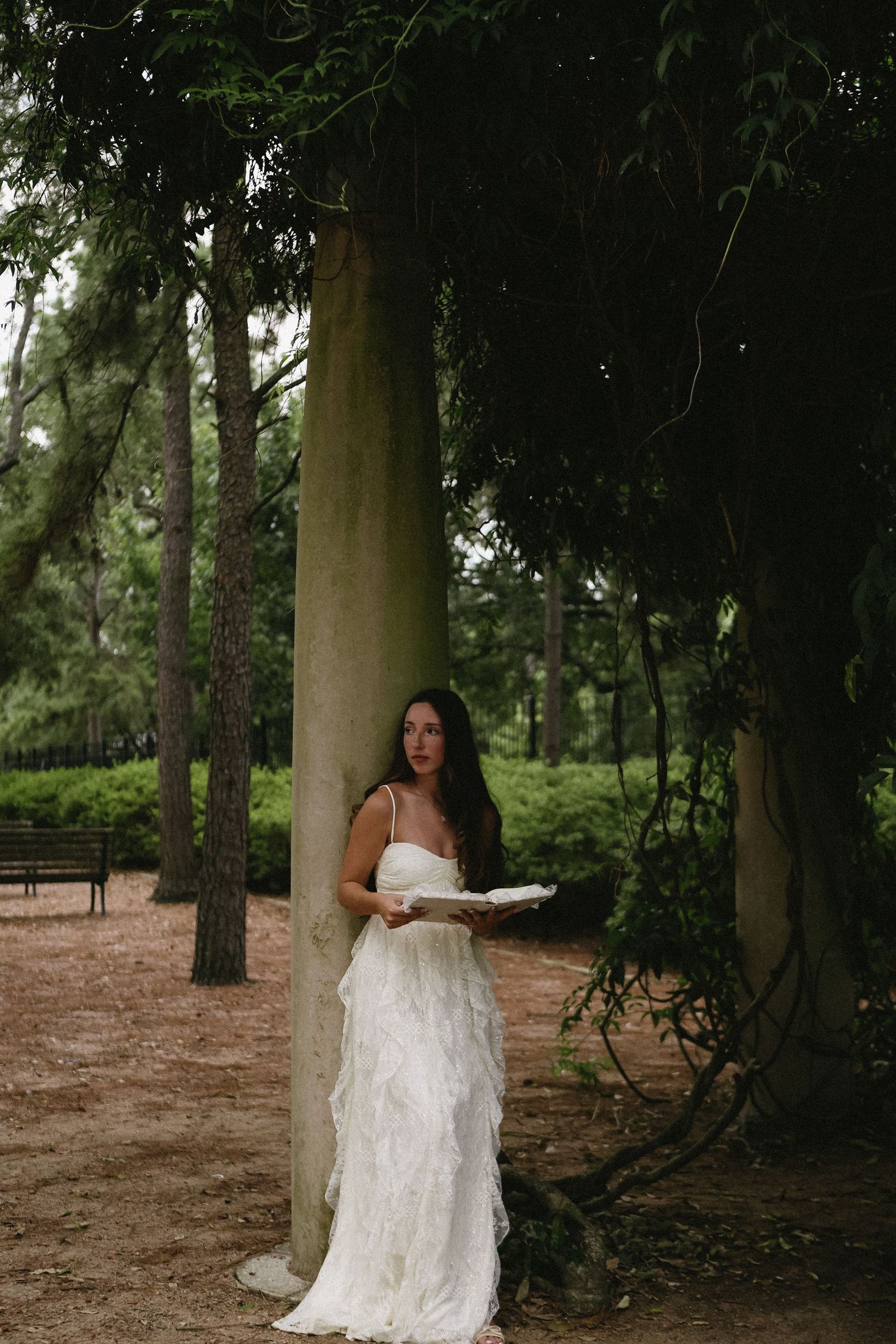 Bride in white ruffled gown standing beside a large tree trunk in a lush green forest setting.
