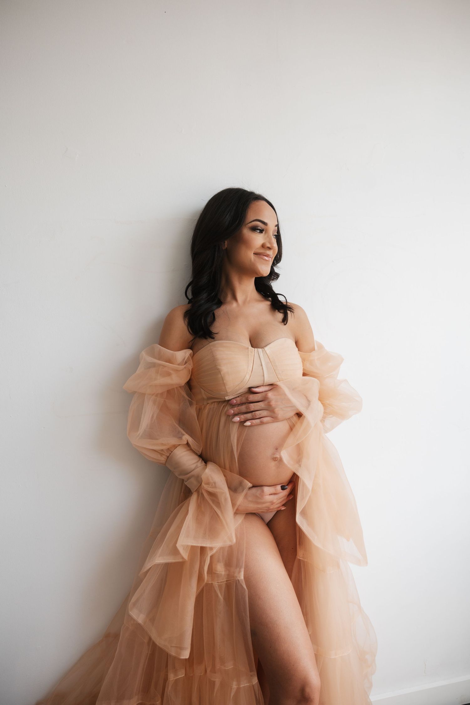 Ethereal maternity photoshoot featuring a sheer peach ruffled tulle robe against white backdrop in elegant poses.