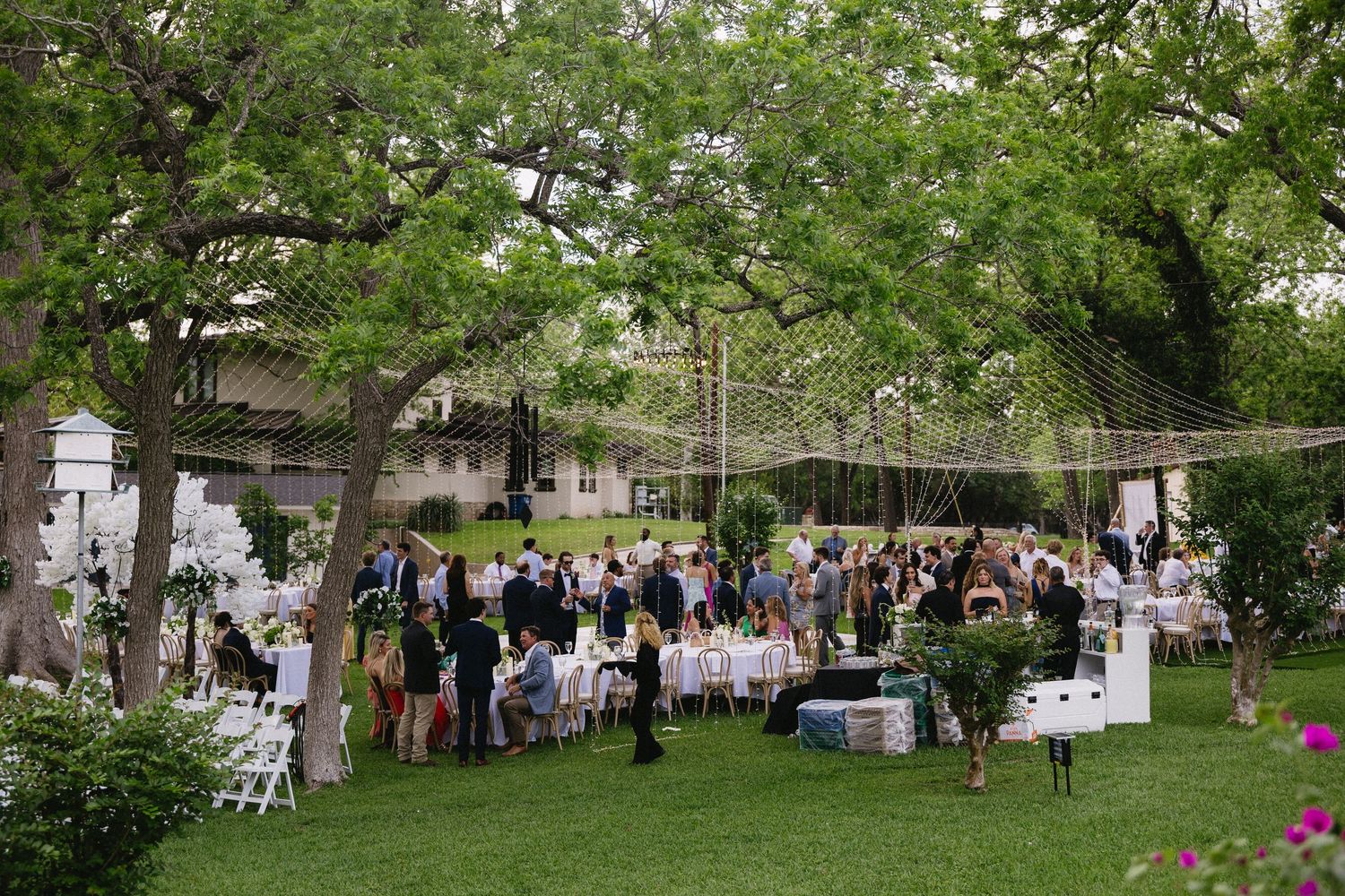 Outdoor garden wedding reception with guests dining under string lights and lush green trees on a sunny day.
