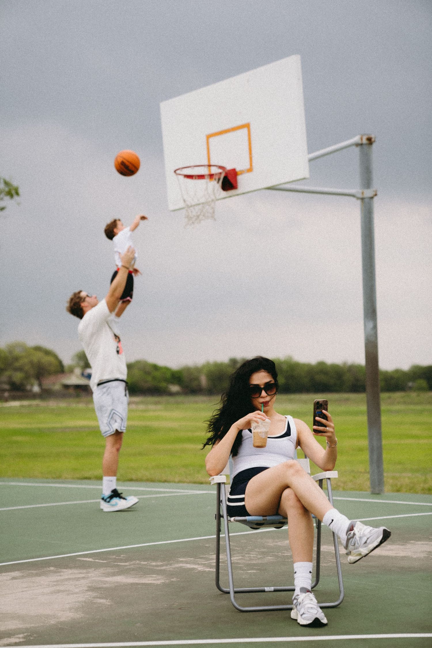 Woman sitting in folding chair using phone on outdoor basketball court while man shoots hoops in background.
