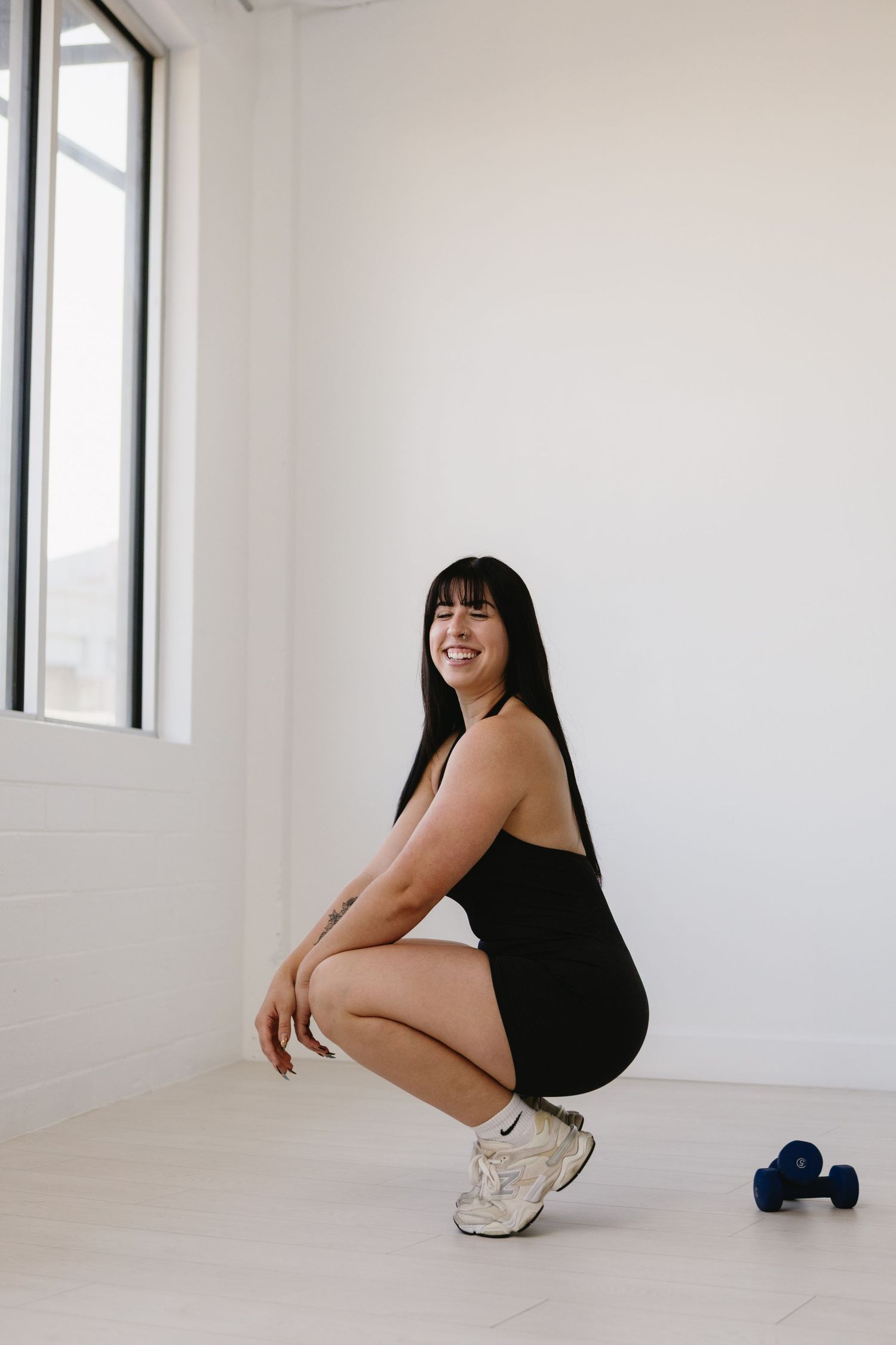 Woman in black bodysuit squatting on white studio floor, smiling, with blue dumbbells nearby.