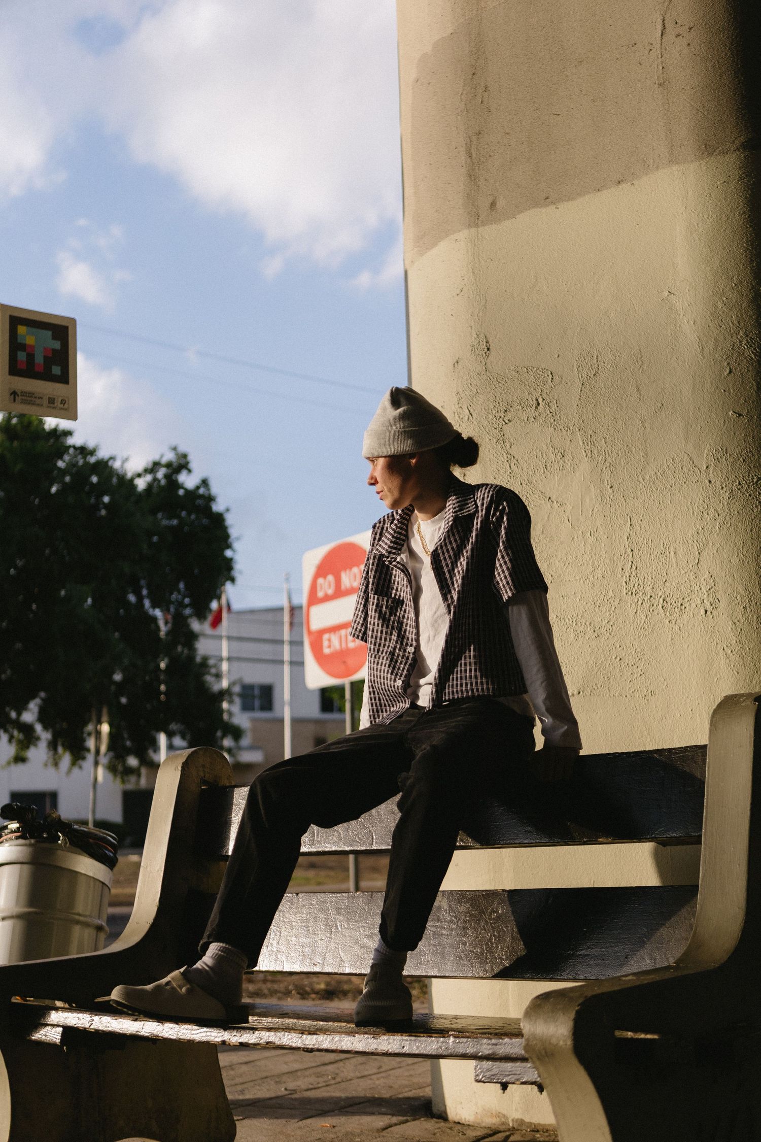 Young person in plaid vest and beanie sits on urban bench beside a wall, with a stop sign and blue sky behind them.