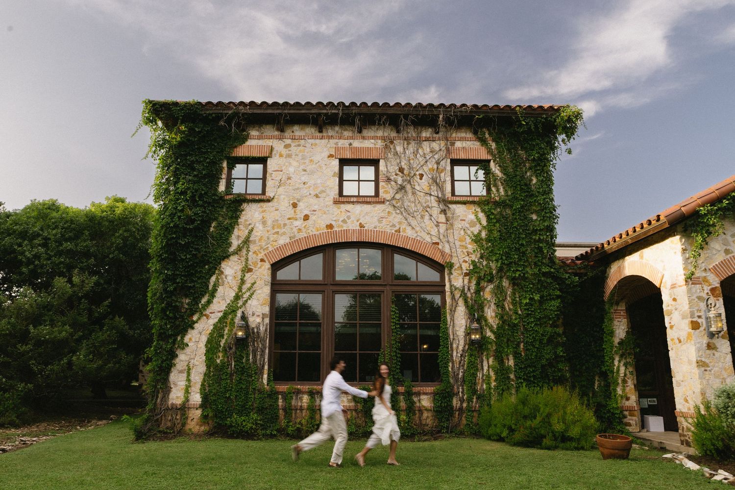 Couple holding hands walking in front of ivy-covered stone villa with arched windows at golden hour.