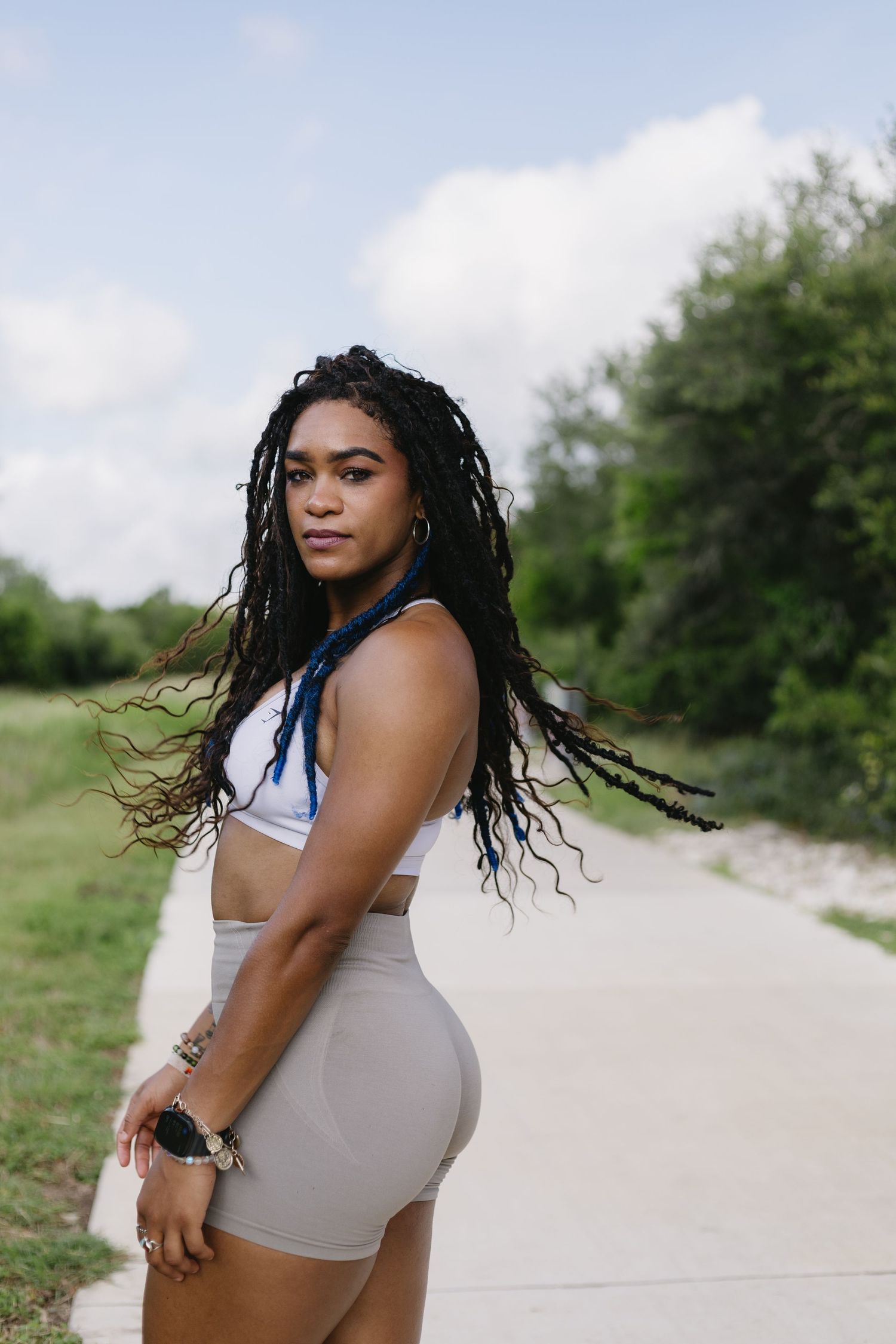 Someone in a sleeveless white top poses gracefully on a path surrounded by green foliage on a cloudy day.