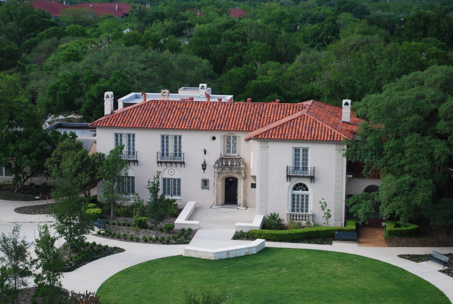 Aerial view of a grand Spanish Colonial Revival mansion with red tile roof and manicured grounds surrounded by lush trees.