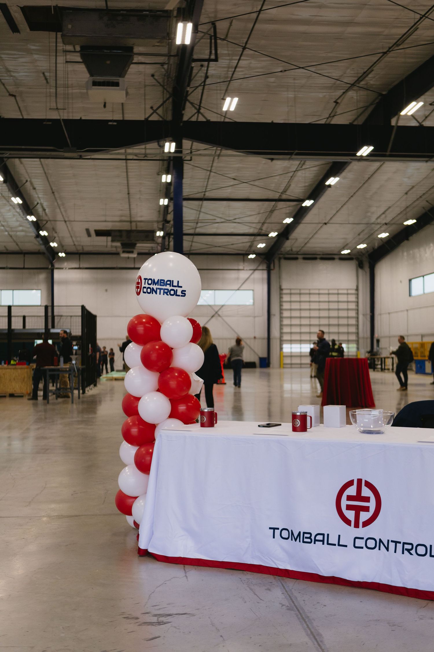 A corporate event setup with red and white balloon decorations and a Tomball Controls branded table display in a warehouse space.