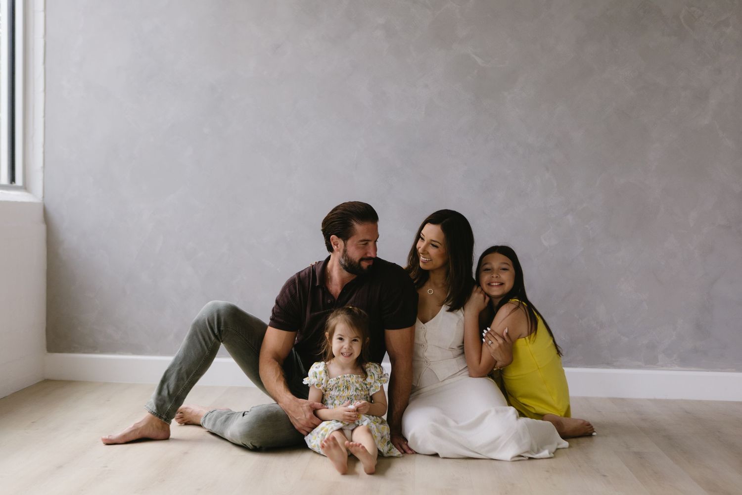 A casual family portrait with parents and children sitting together on a hardwood floor.