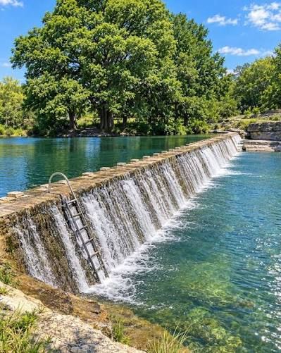 A scenic stone dam waterfall flows into a clear blue-green river surrounded by lush green trees on a sunny day.