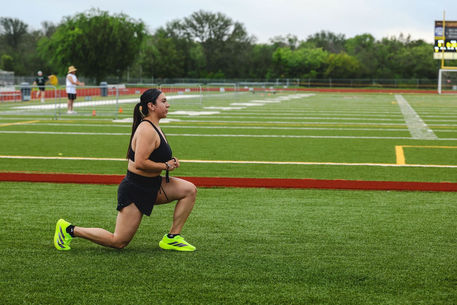 Woman doing lunges on a green turf athletic track field wearing neon yellow sneakers and black athletic wear.