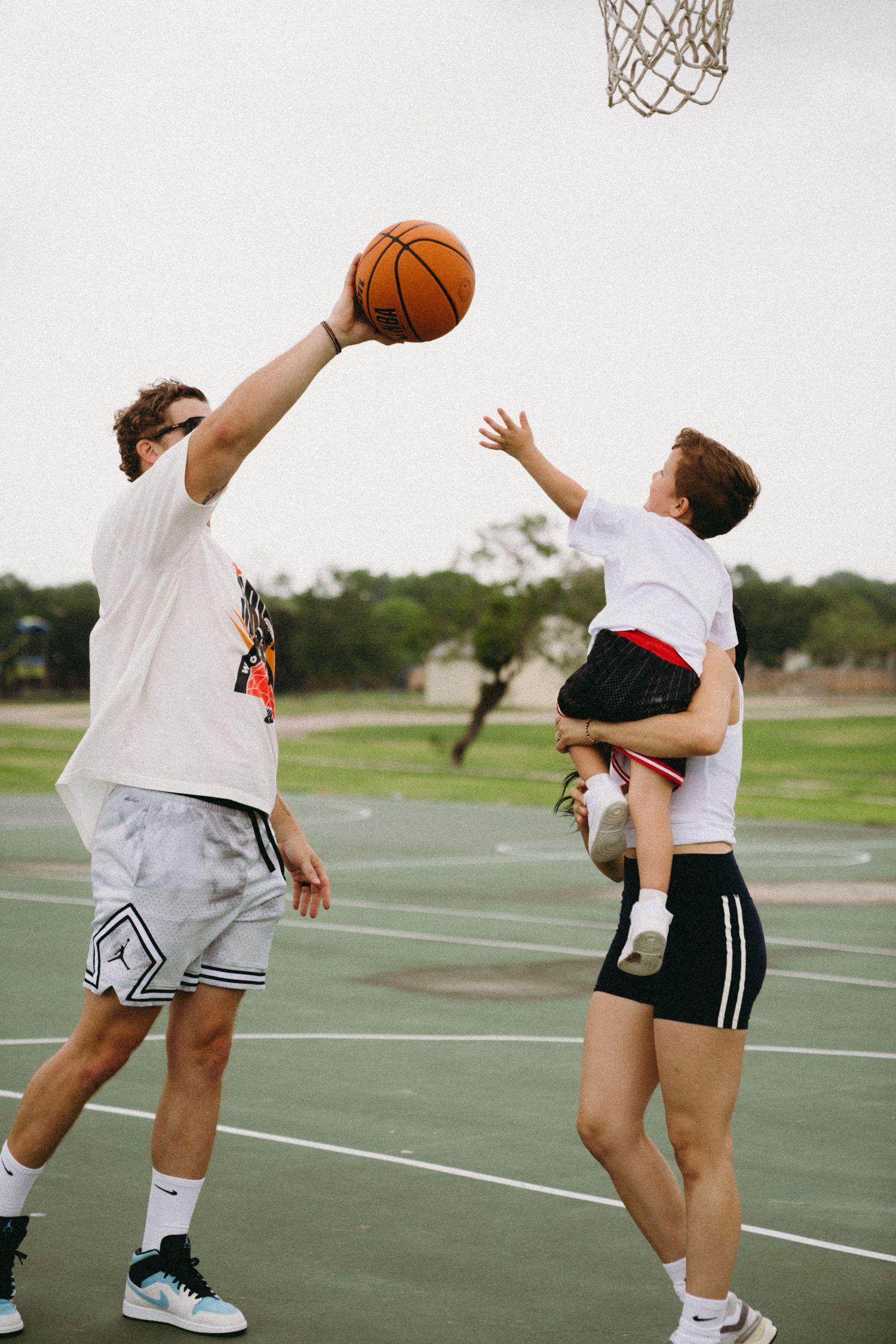 Two men and a child play basketball outdoors, one lifting the child to reach the hoop on a green court.