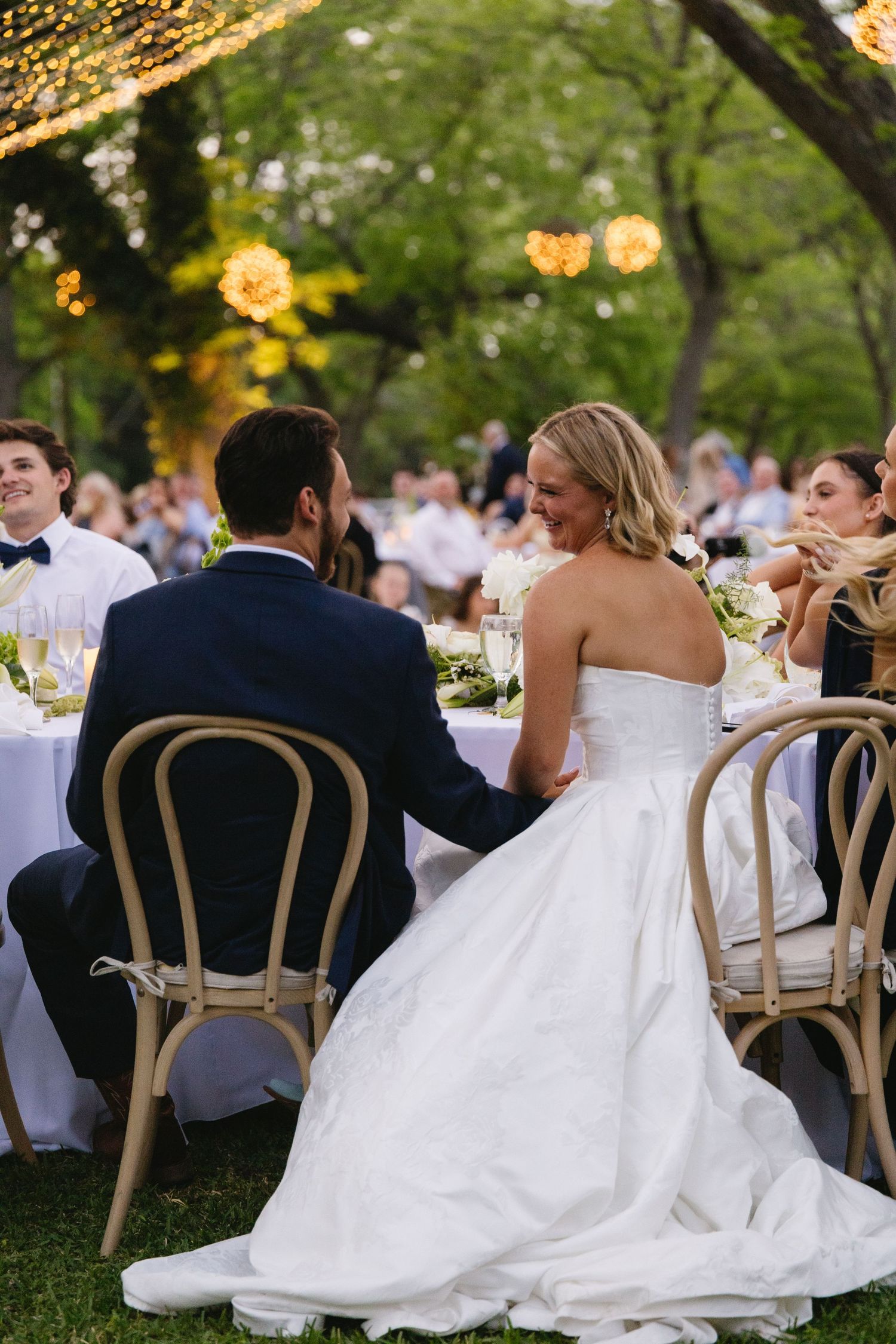 Bride and groom holding hands at outdoor wedding reception dinner under string lights surrounded by guests.