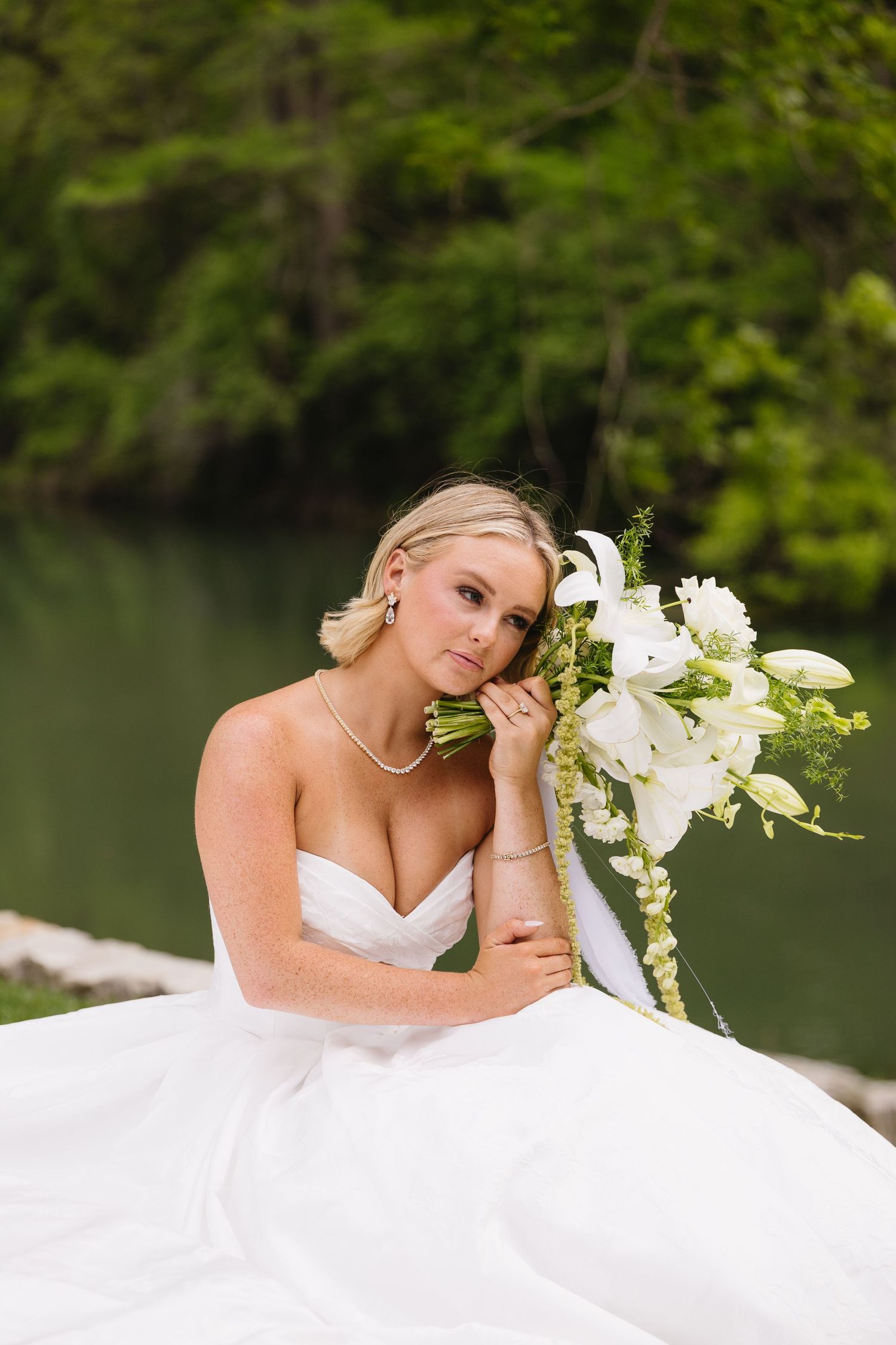 Bride in strapless white gown holds white floral bouquet near serene lake with lush green trees backdrop.