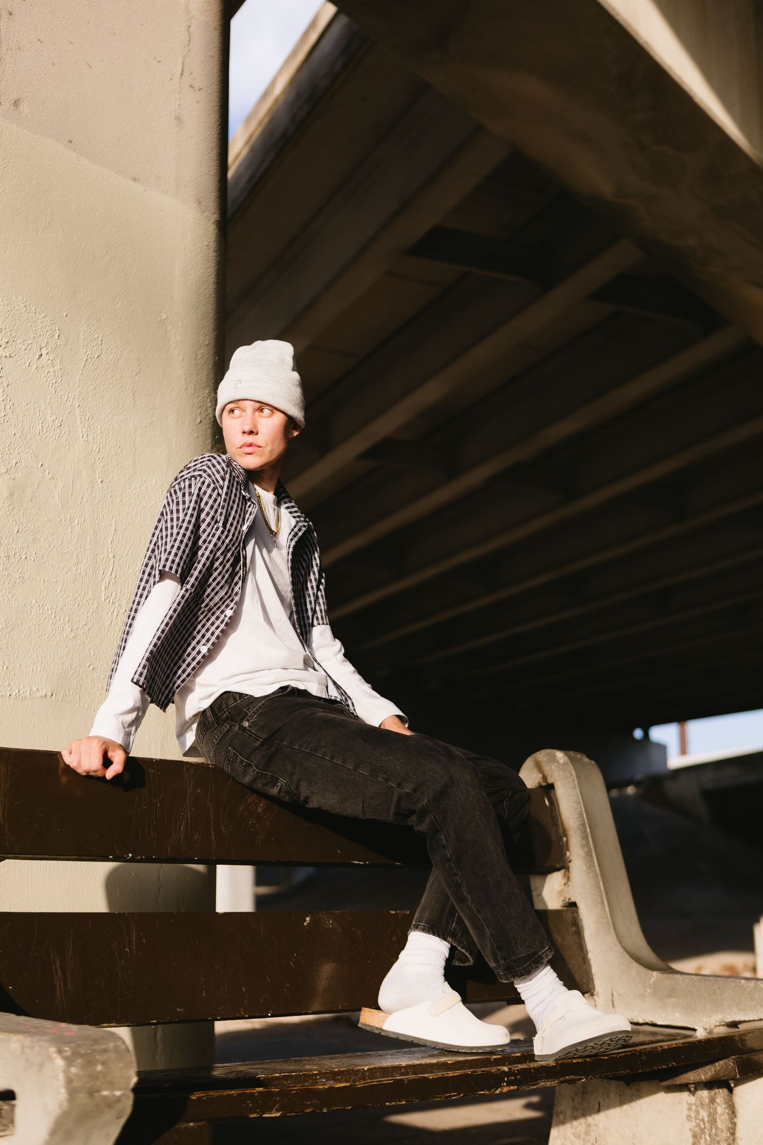 Young man in plaid shirt, beanie hat, and black jeans sitting under a concrete overpass in urban setting.