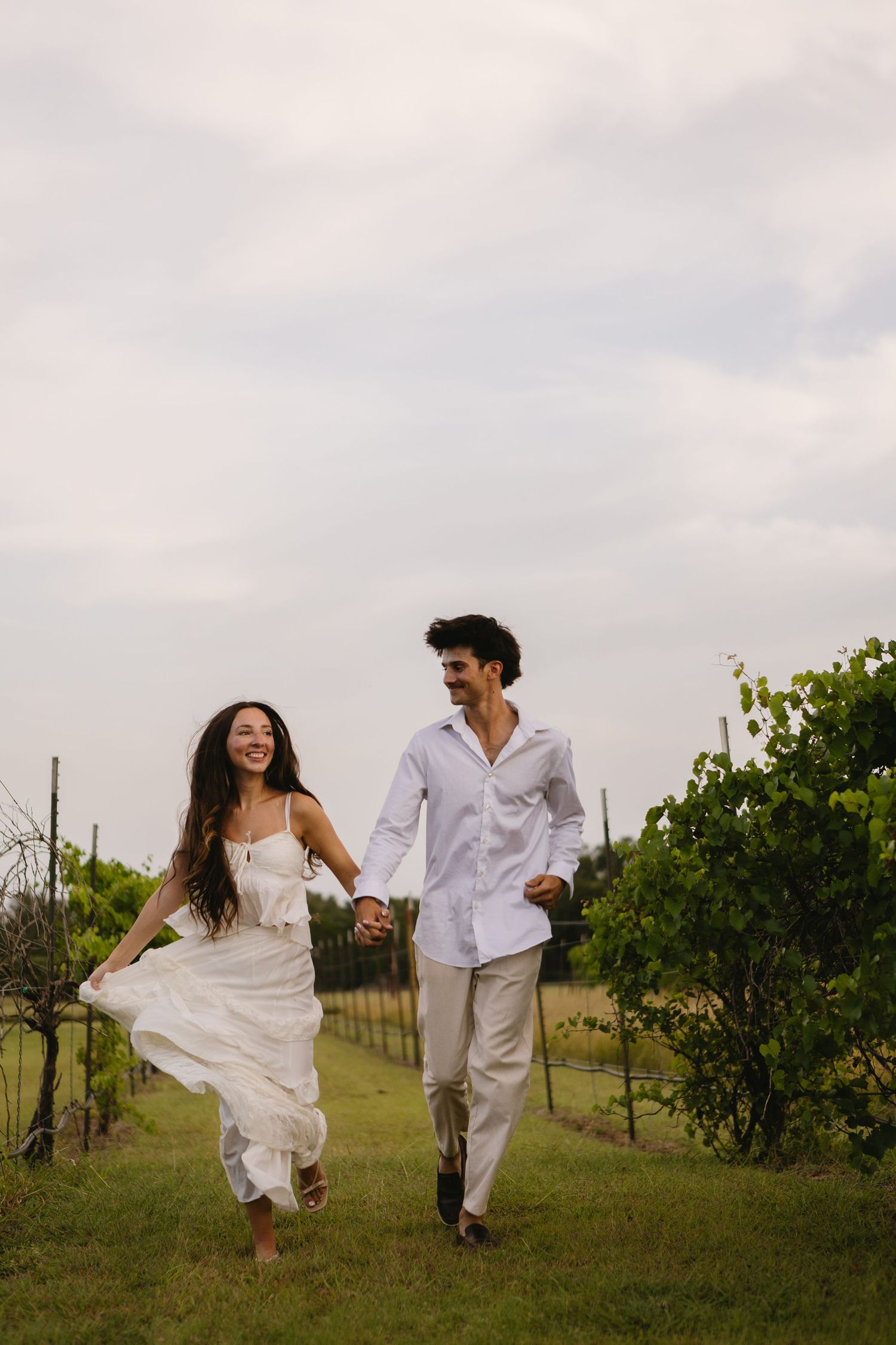 A joyful couple in white outfits holds hands while running through a lush vineyard on a cloudy day.
