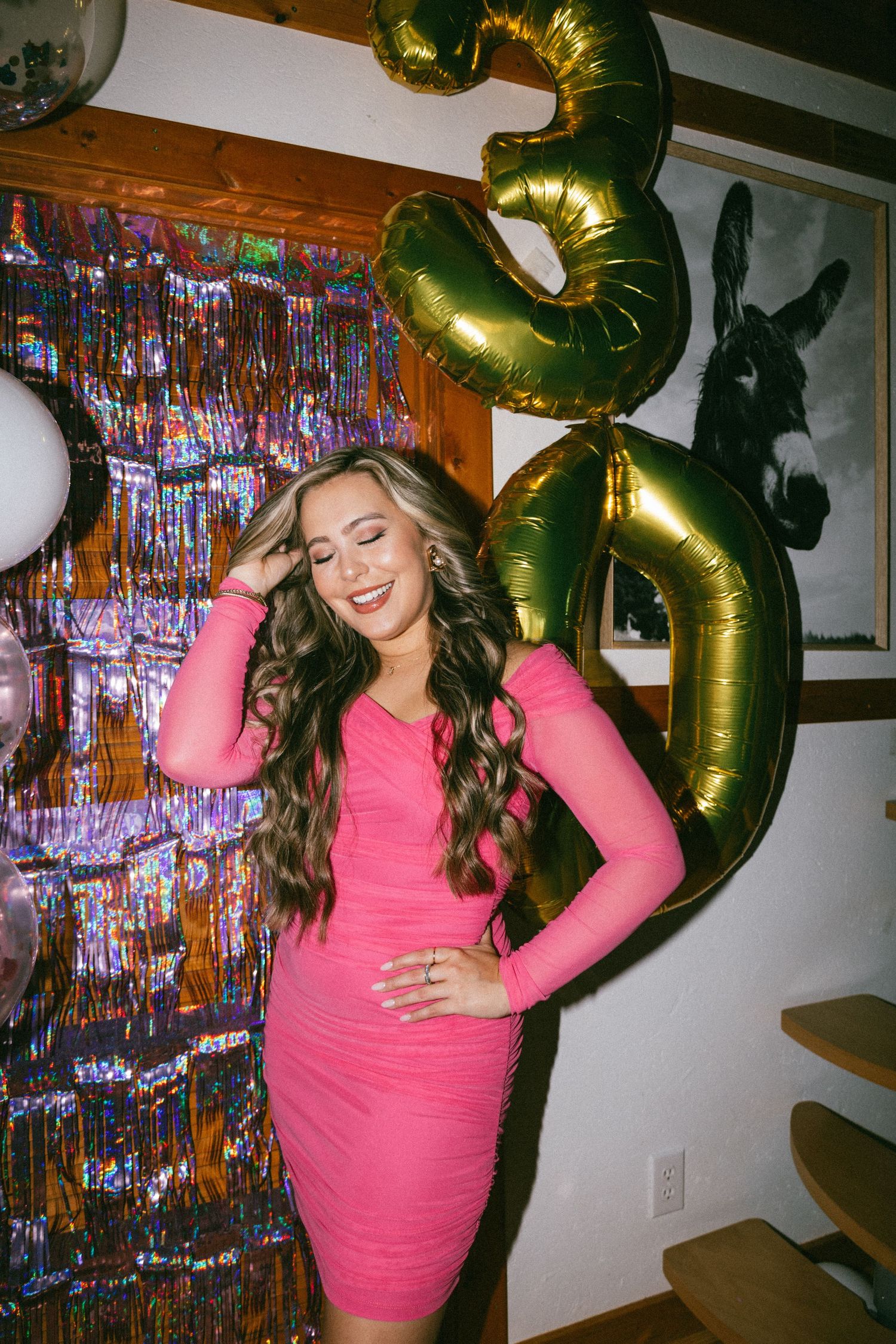 Woman in pink dress posing at party with gold balloon number and sparkly backdrop.