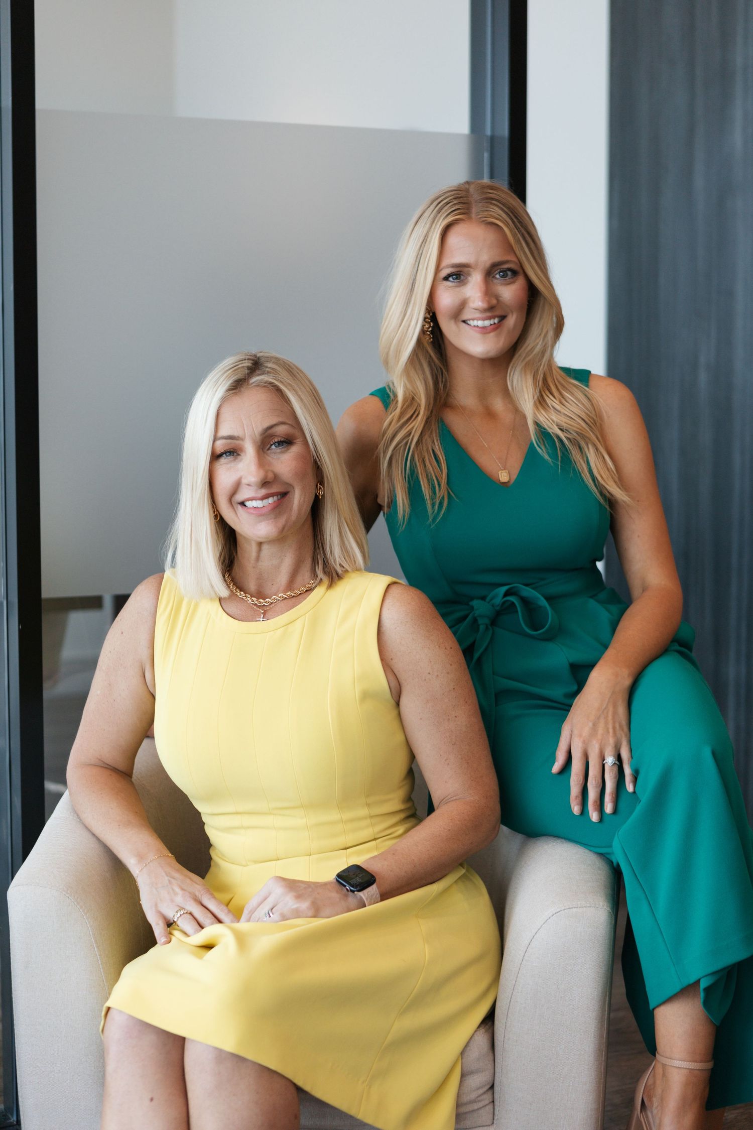 Two women in professional attire, one wearing a yellow sleeveless dress and one in a green jumpsuit, sitting in a modern space.